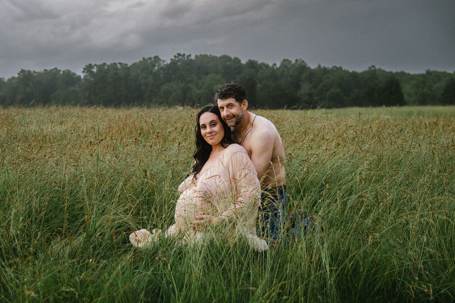 A romantic maternity photoshoot in a lush green field at sunset with dark storm clouds overhead.
