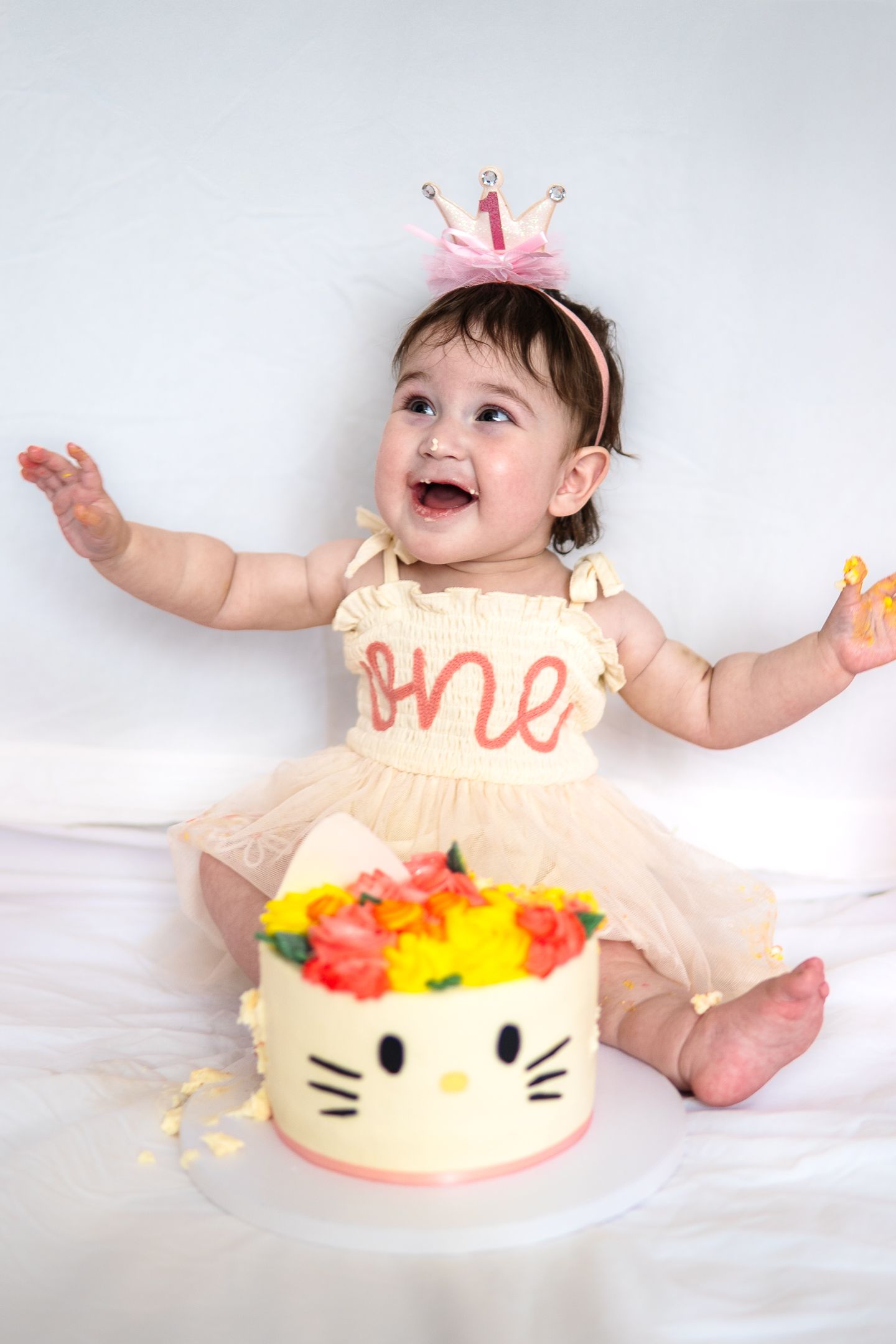 Happy baby in cream dress and pink crown sits with colorful Hello Kitty themed first birthday cake on white background.