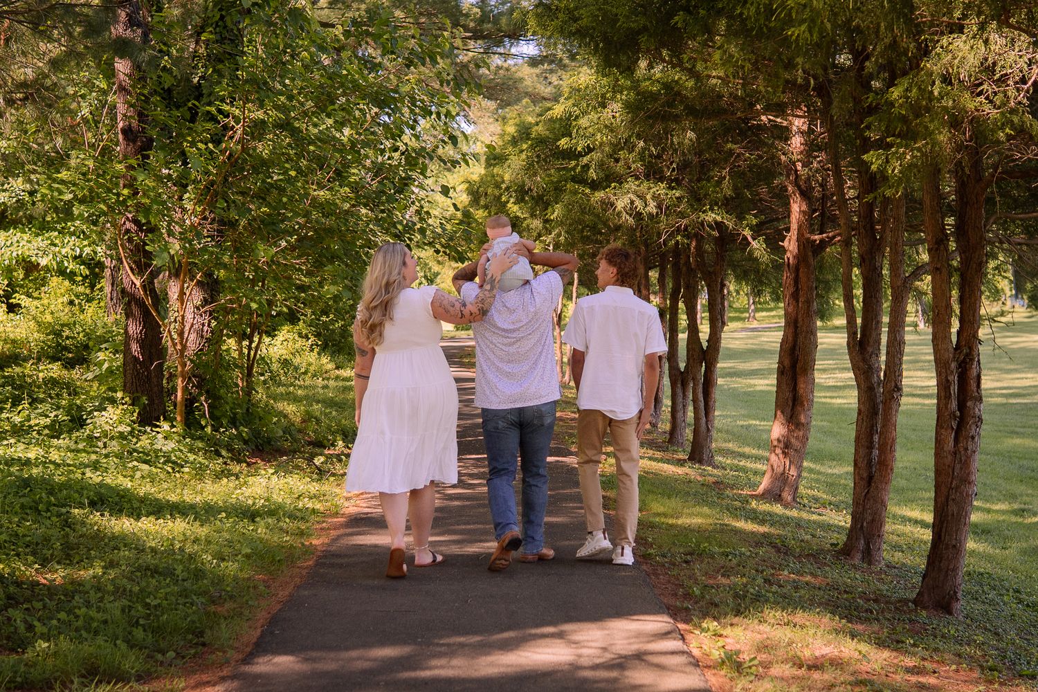 Three people walk together down a tree-lined paved path on a sunny summer afternoon in a park.