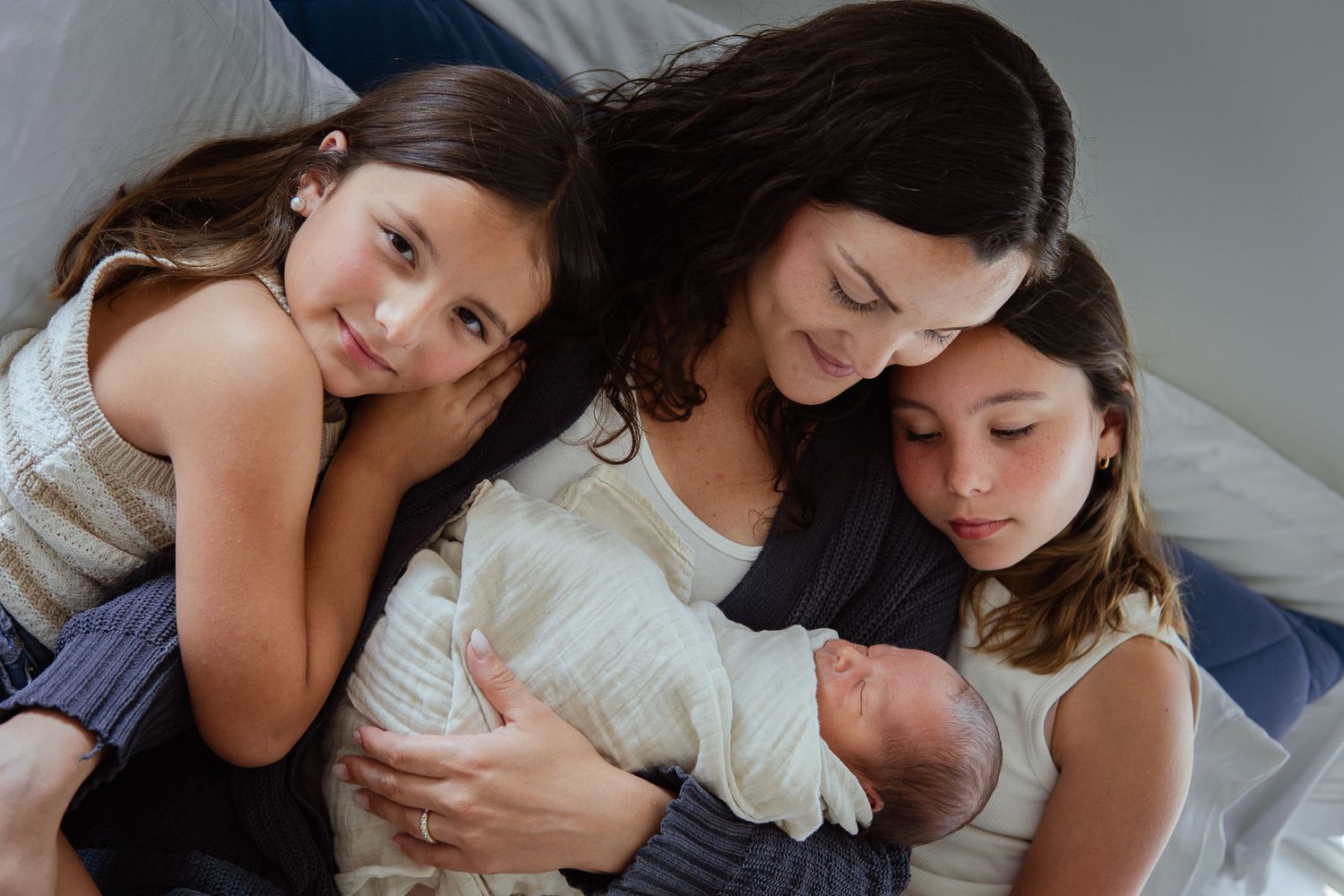 Intimate family portrait of siblings gathering around newborn baby on bed.