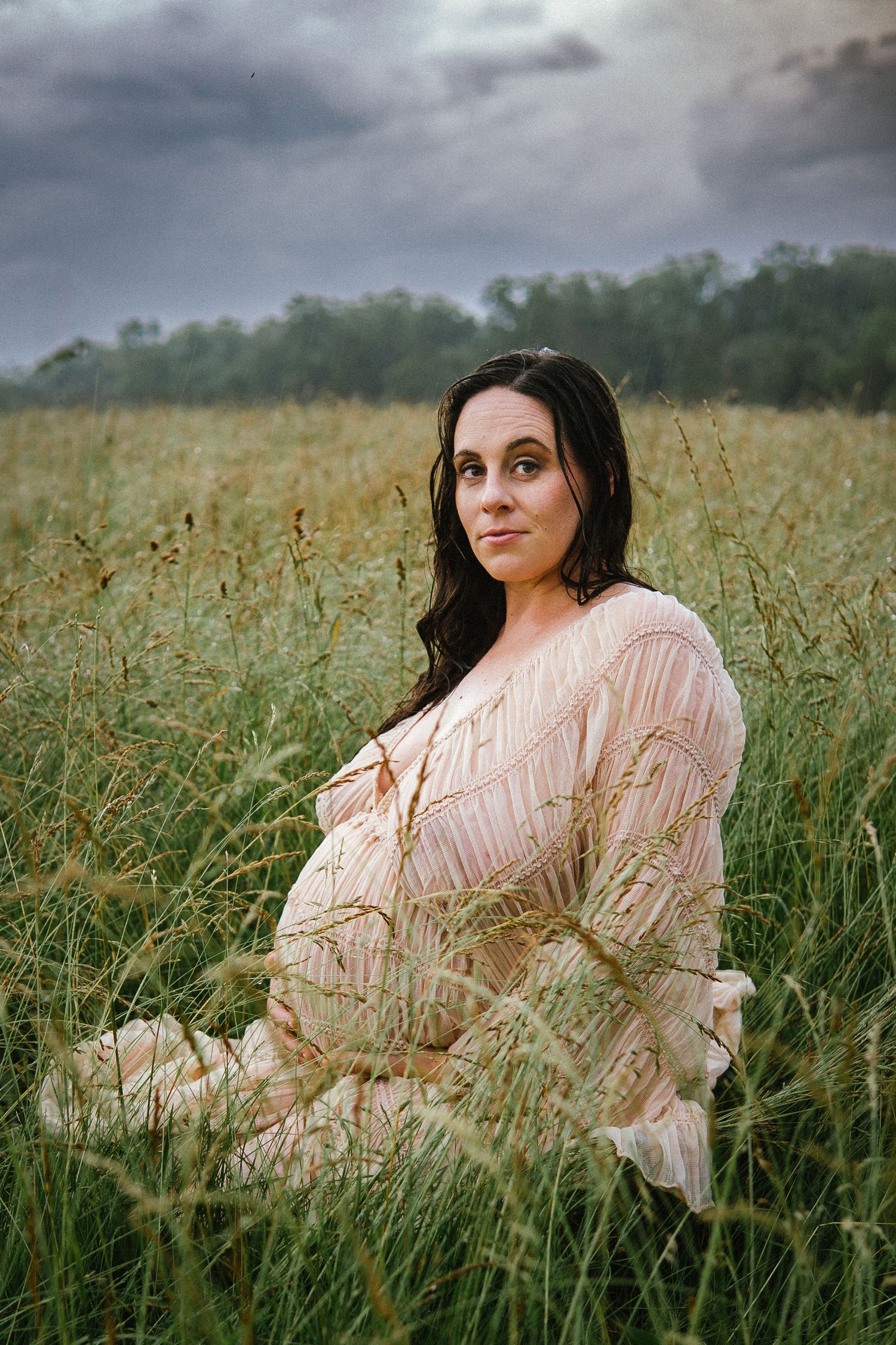 A woman in a beige dress sits thoughtfully in a field of tall grass under moody storm clouds.