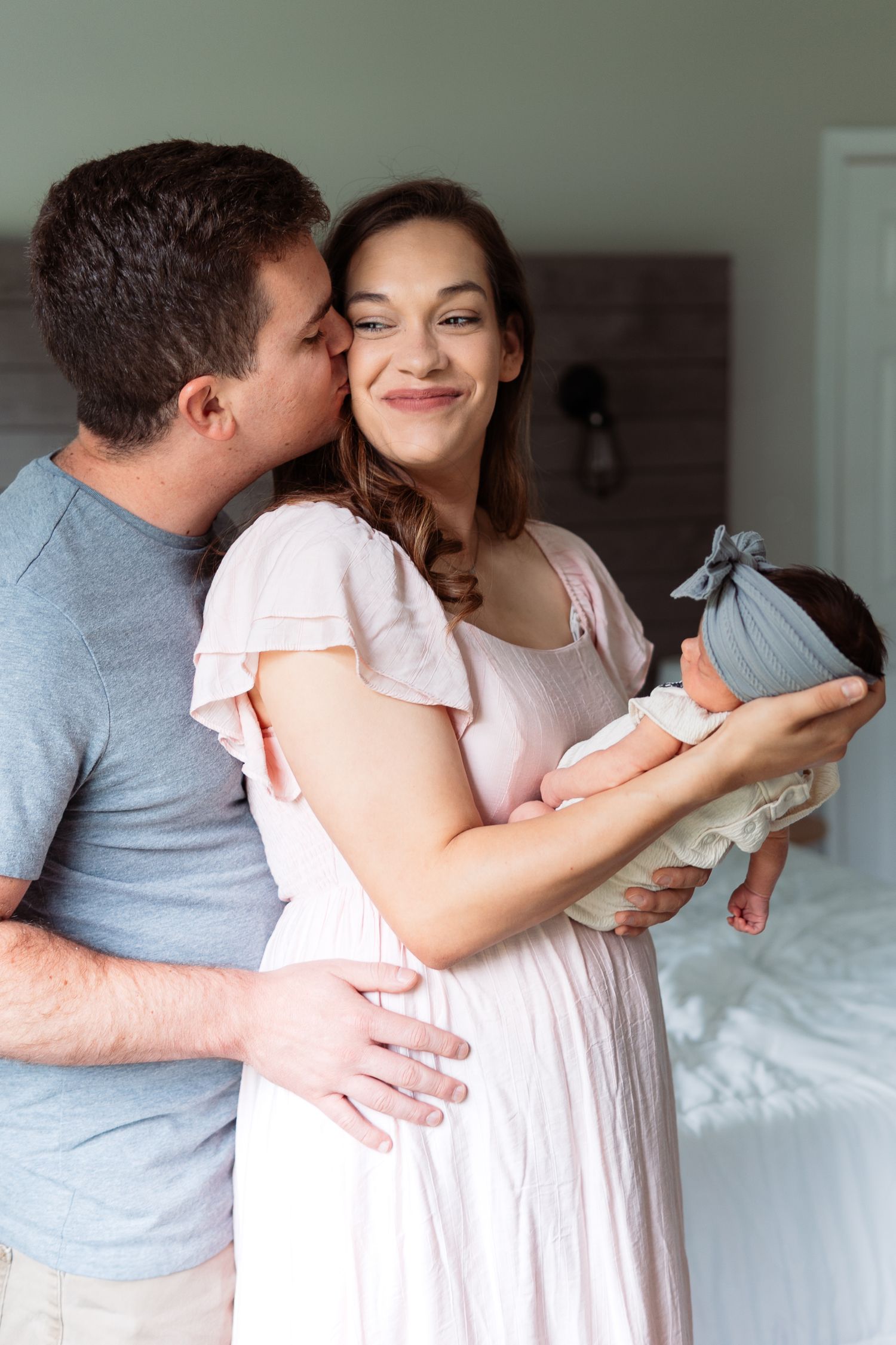 A sweet moment between a couple holding their newborn baby wrapped in gray swaddling cloth in a bright room.