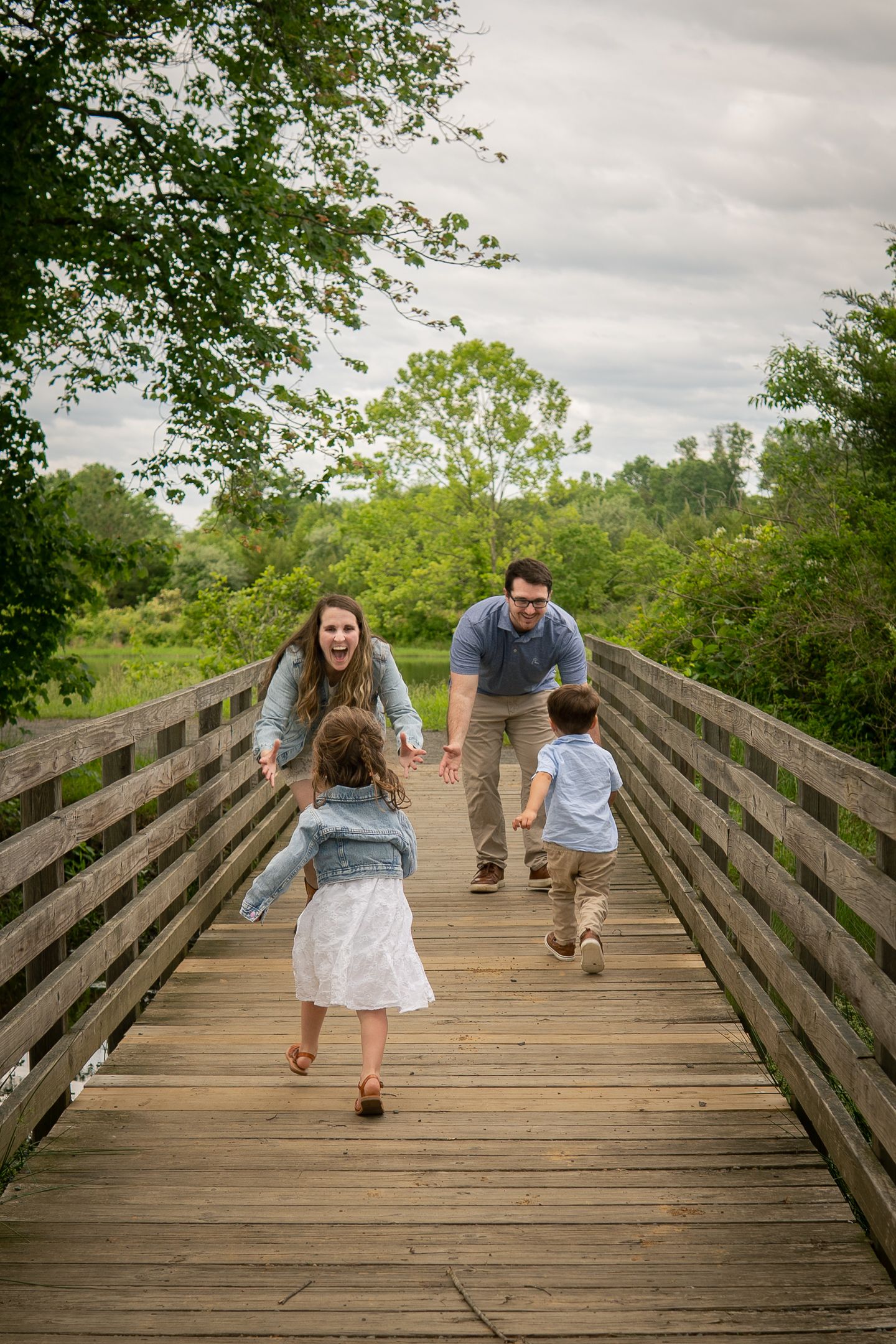 Family playing together on a wooden footbridge in a lush green park setting during an overcast day.