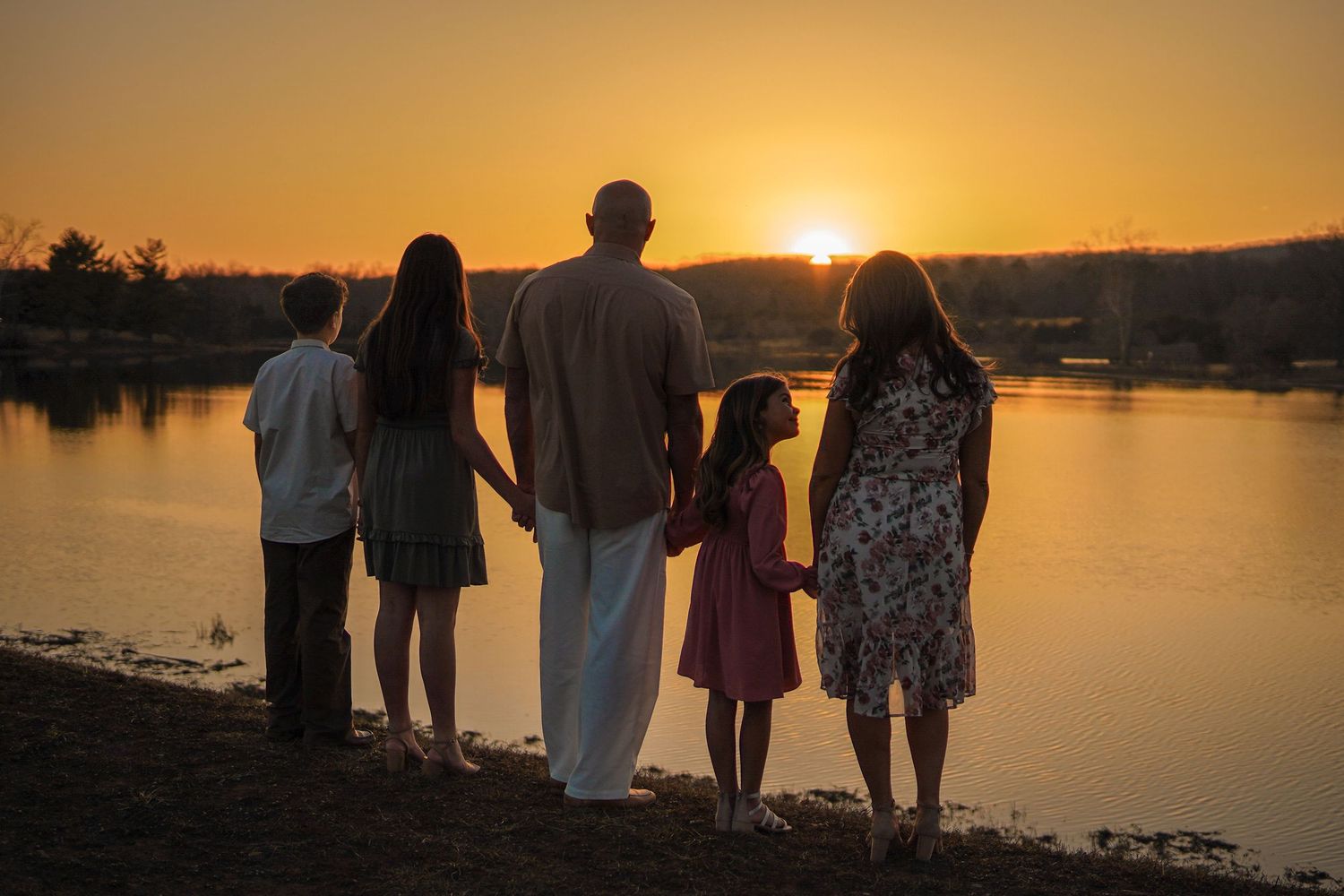 Silhouettes of a family watch the sun setting over a tranquil lake, creating a peaceful golden hour scene.