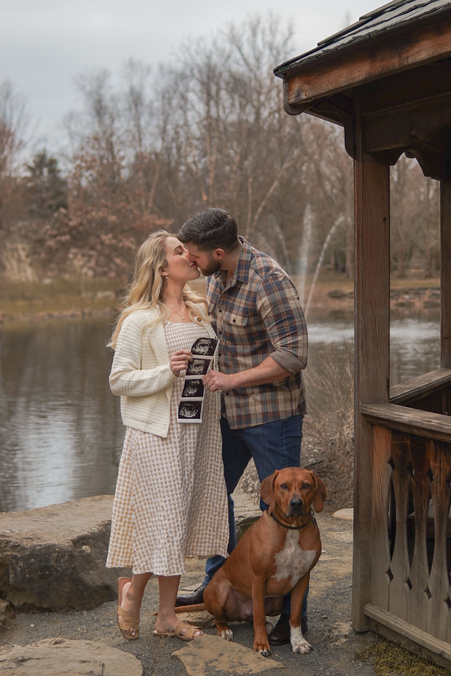 A couple shares a tender moment by a lake with their brown dog while holding ultrasound photos near a wooden gazebo.