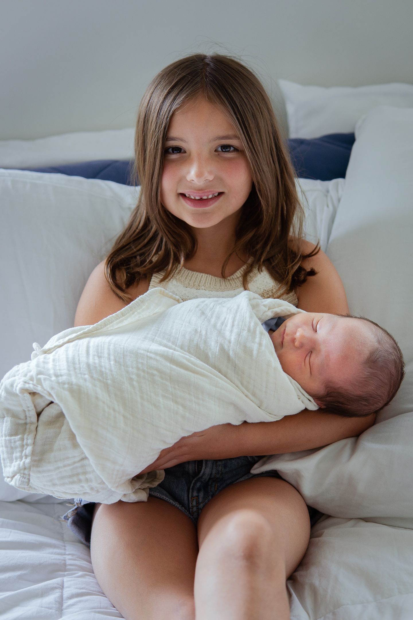 Lifestyle photos of siblings bonding with newborn on white bedding.