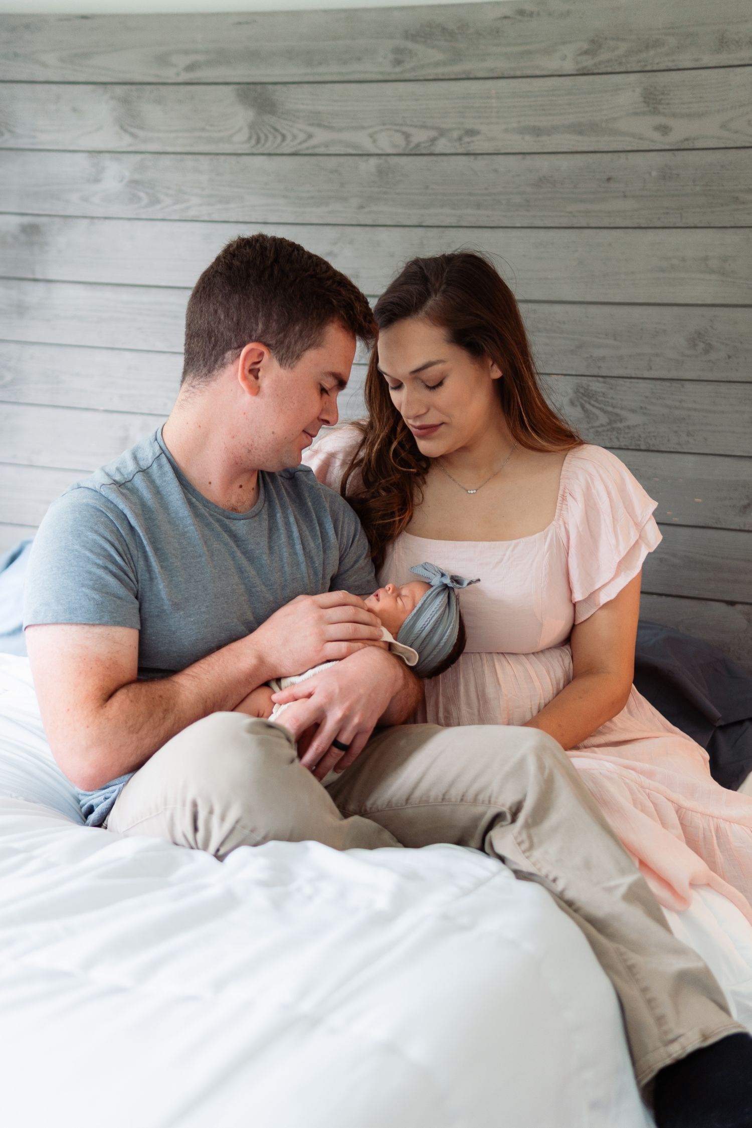 Two people sit together on a bed with white sheets against a rustic wooden wall background.
