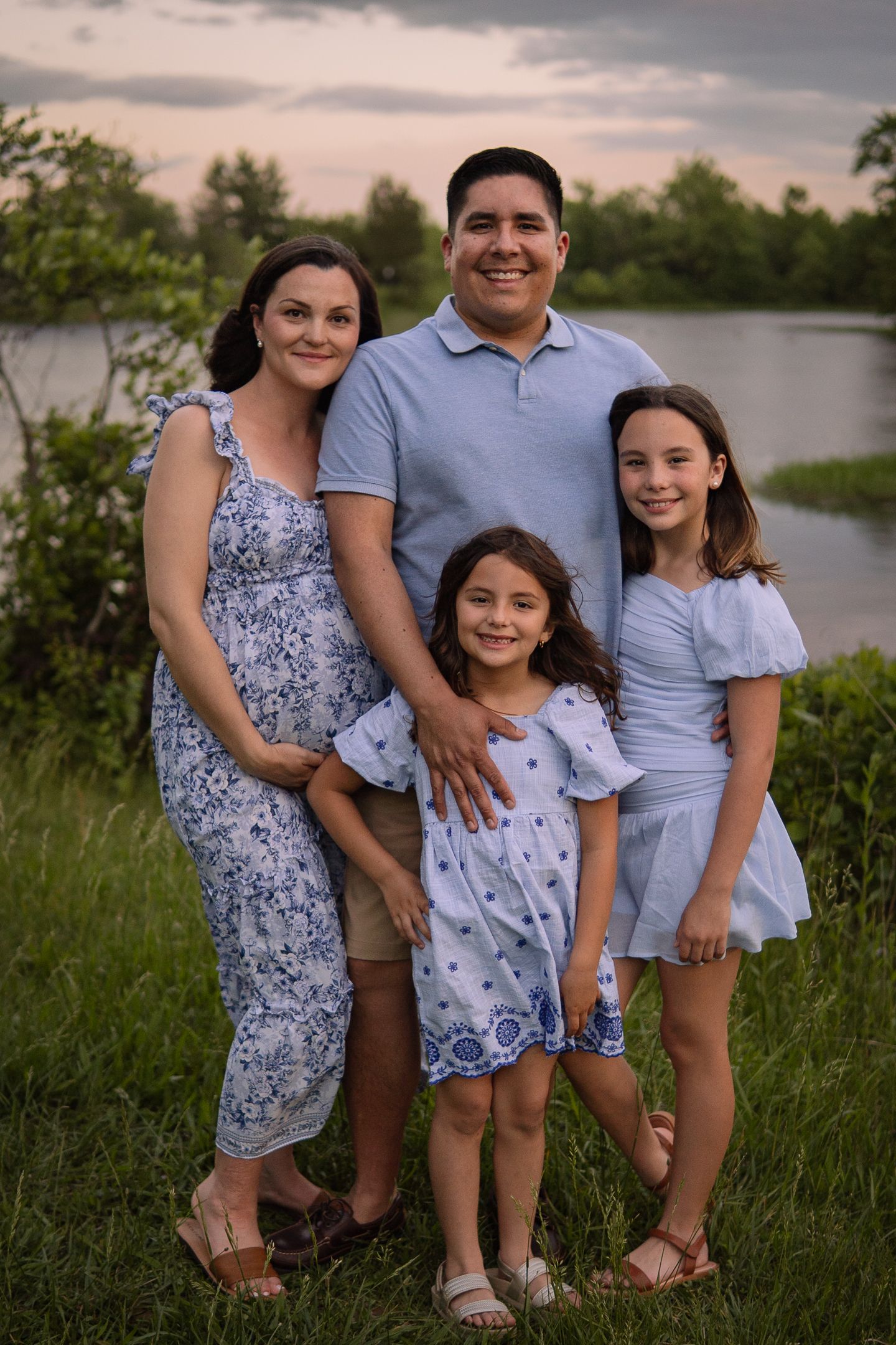Family portrait with coordinated outfits posing together by water at dusk.