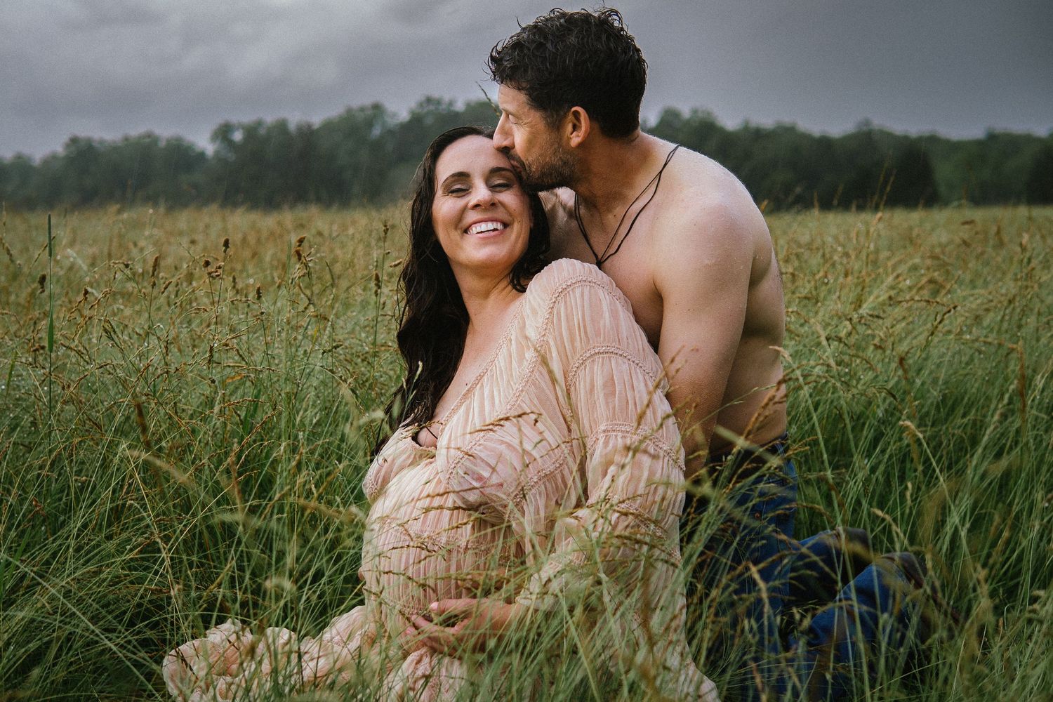 A couple embraces intimately in a tall grass field during a romantic outdoor photoshoot at sunset.