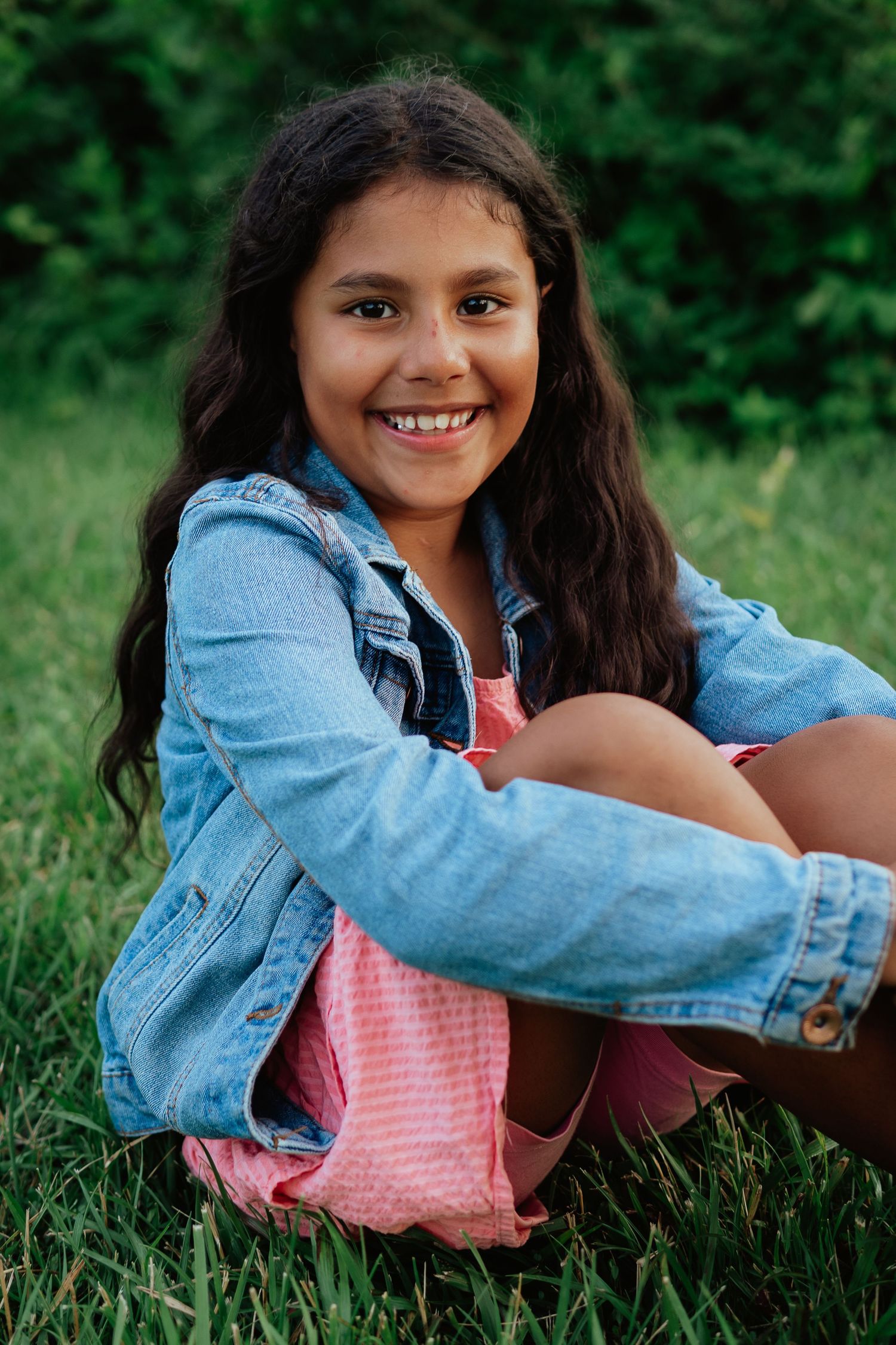 A young person in a denim jacket and pink dress sits on grass, smiling brightly at the camera.