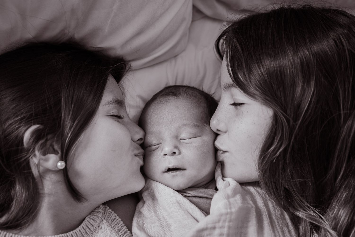 A black and white portrait of two parents giving gentle kisses to their sleeping newborn baby.