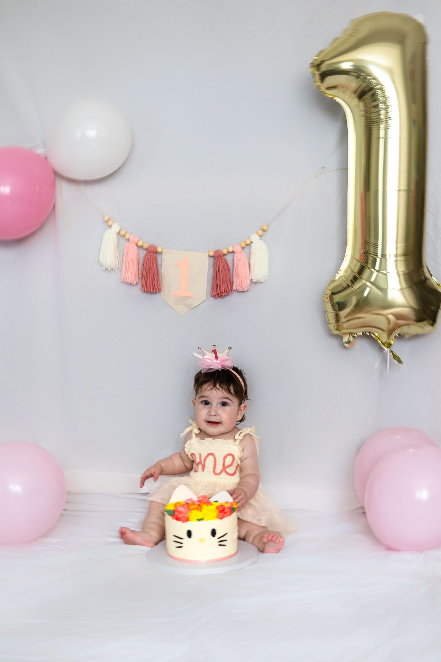 First birthday celebration setup with gold number one balloon, pink decorations and Hello Kitty cake against white backdrop.