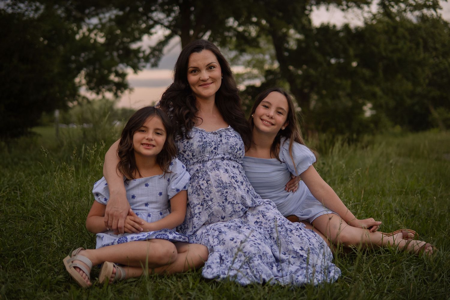 Three smiling people sit together on grass wearing coordinated purple and blue outfits at sunset.