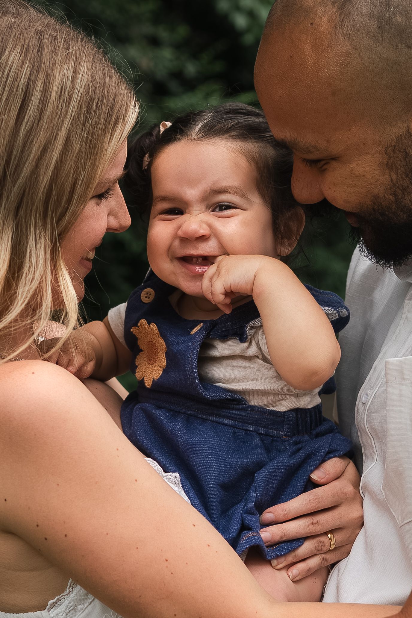 A joyful toddler in blue overalls smiles while being held by parents in an outdoor setting.