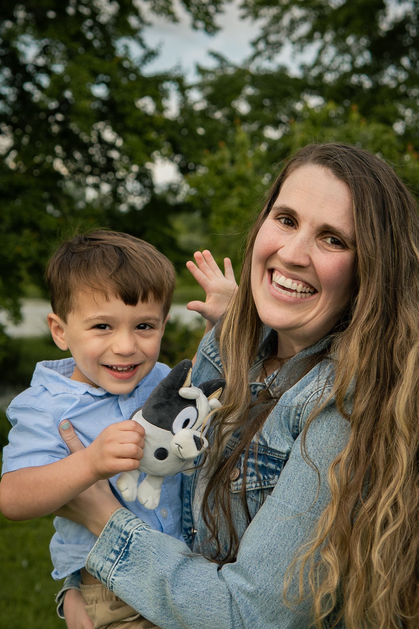 A joyful outdoor portrait featuring long wavy hair, denim jacket, and a small stuffed animal toy in a natural setting.