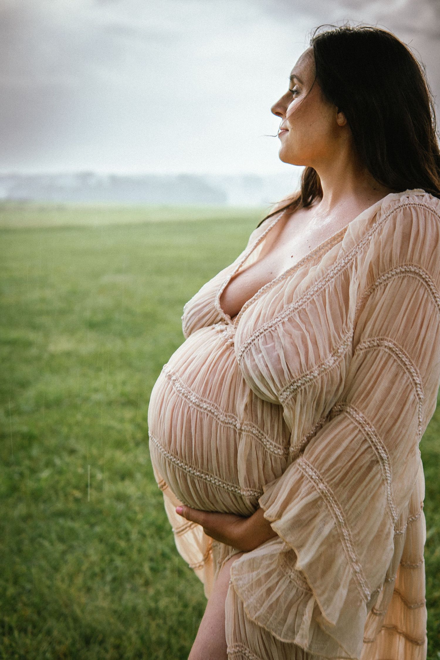 An expectant mother in a flowy pink dress stands in profile against a misty green field landscape.