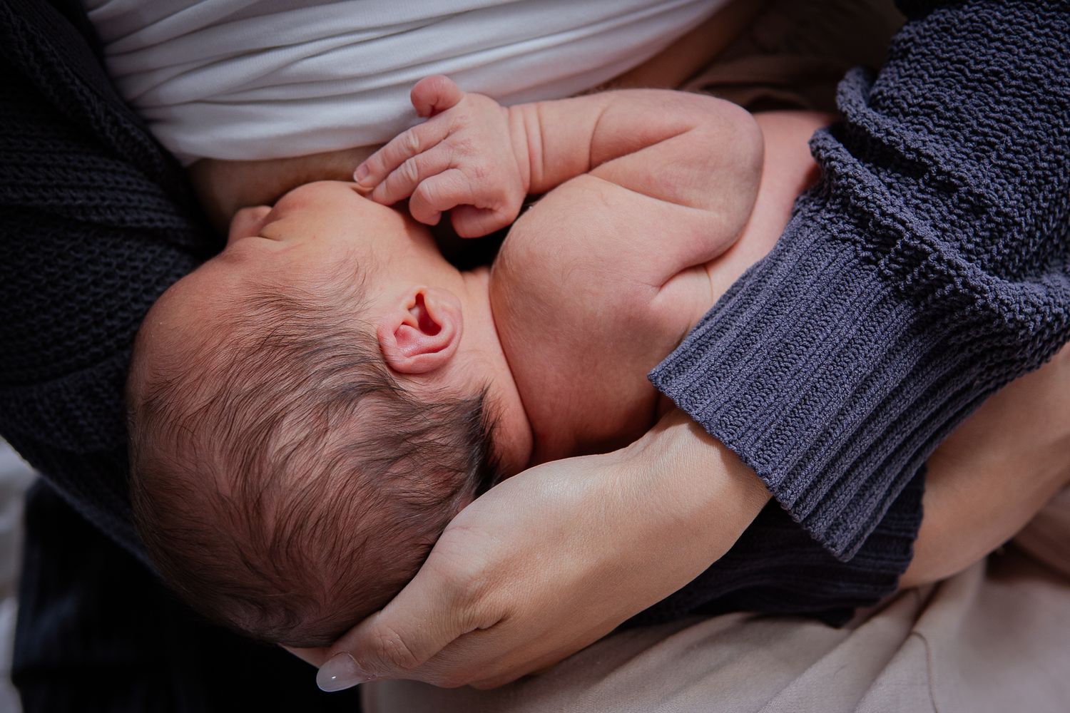 Sleeping newborn nestled in parent's arms with dark knit sweater.