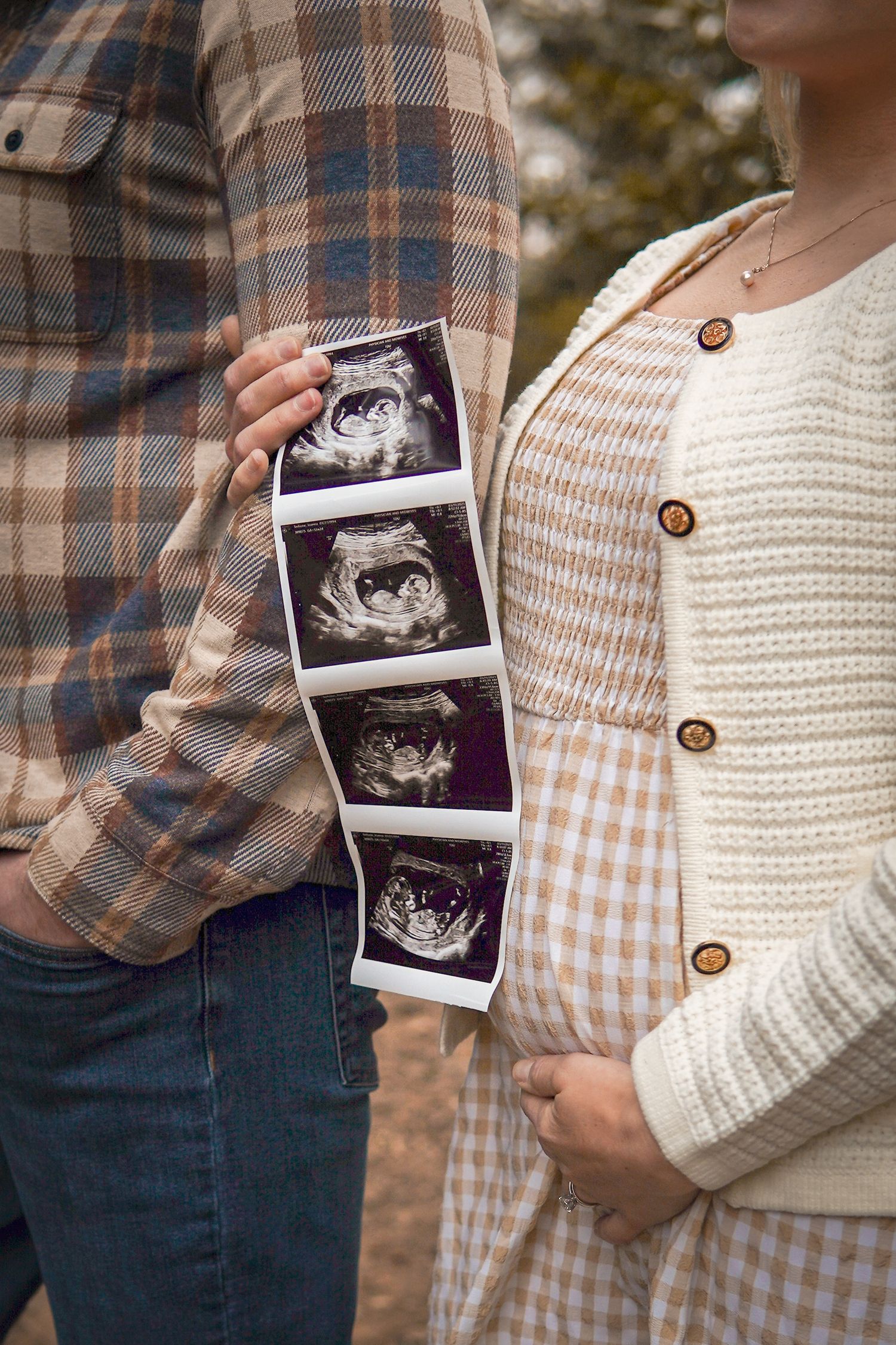 Couple holding ultrasound photos showing pregnancy announcement while wearing plaid shirt and white cardigan outdoors.
