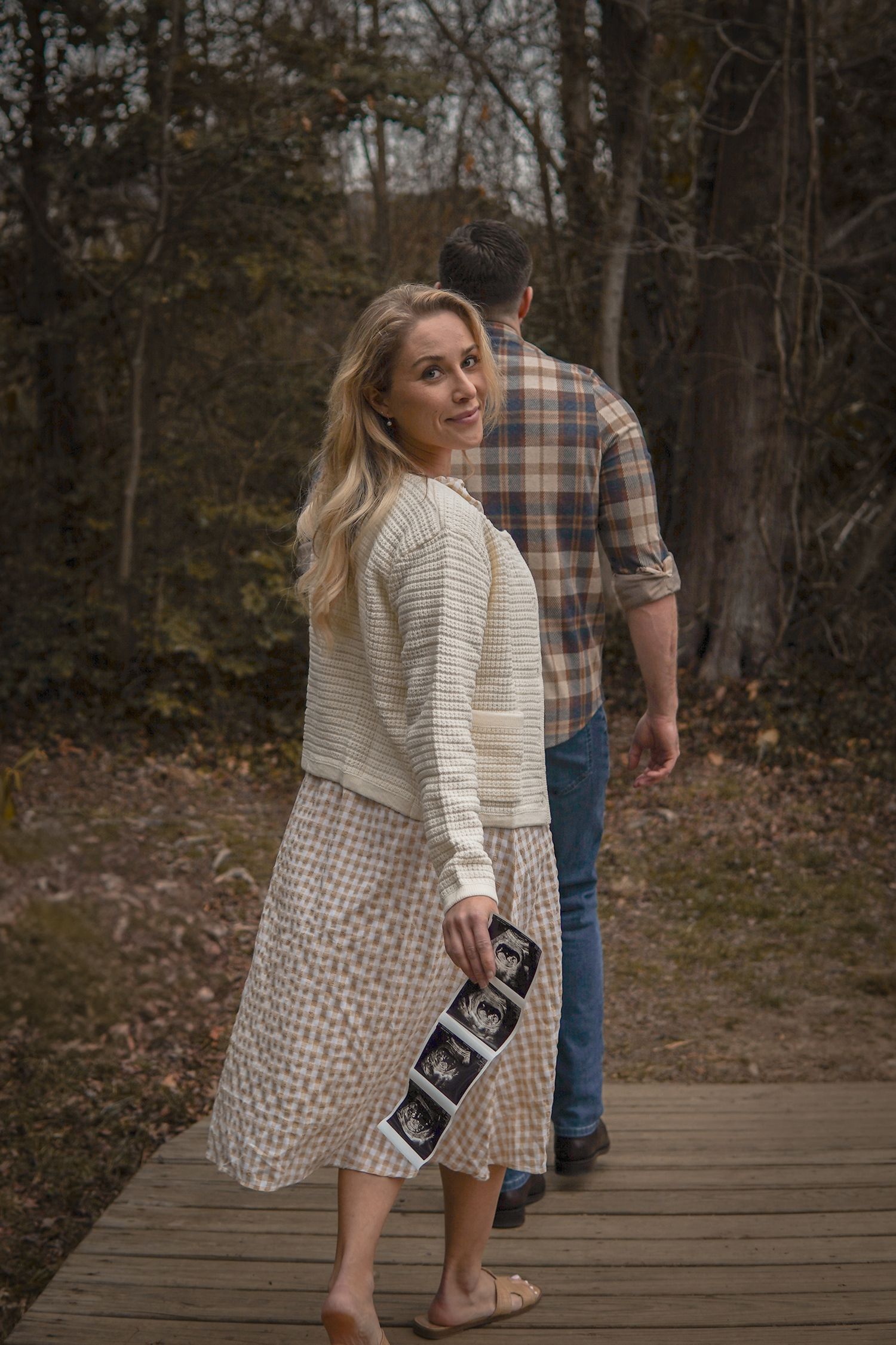 A couple walks along a wooden boardwalk path through autumn woods while holding sonogram photos.