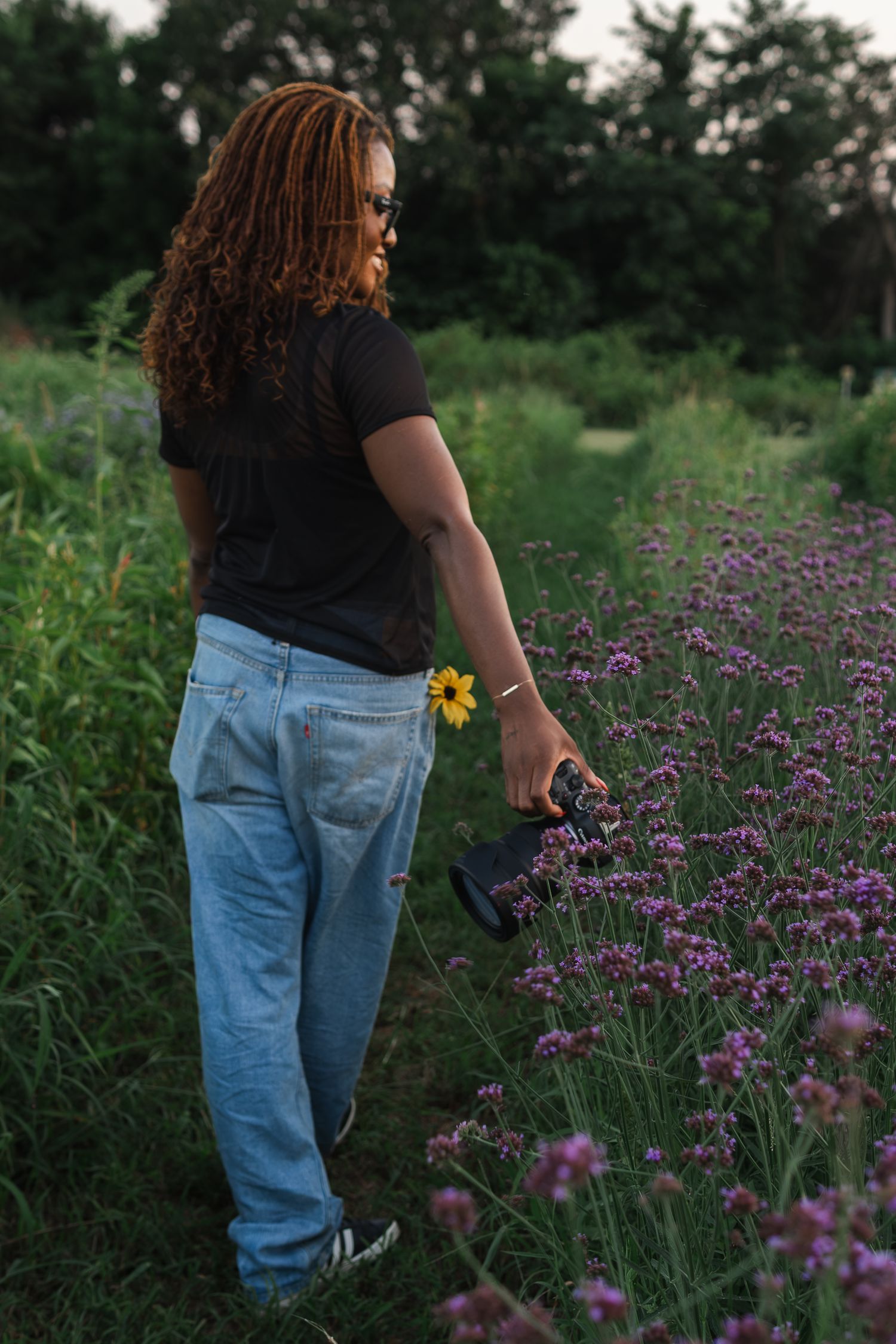 Someone holding a camera stands in a field of purple wildflowers at sunset wearing jeans and a black t-shirt.