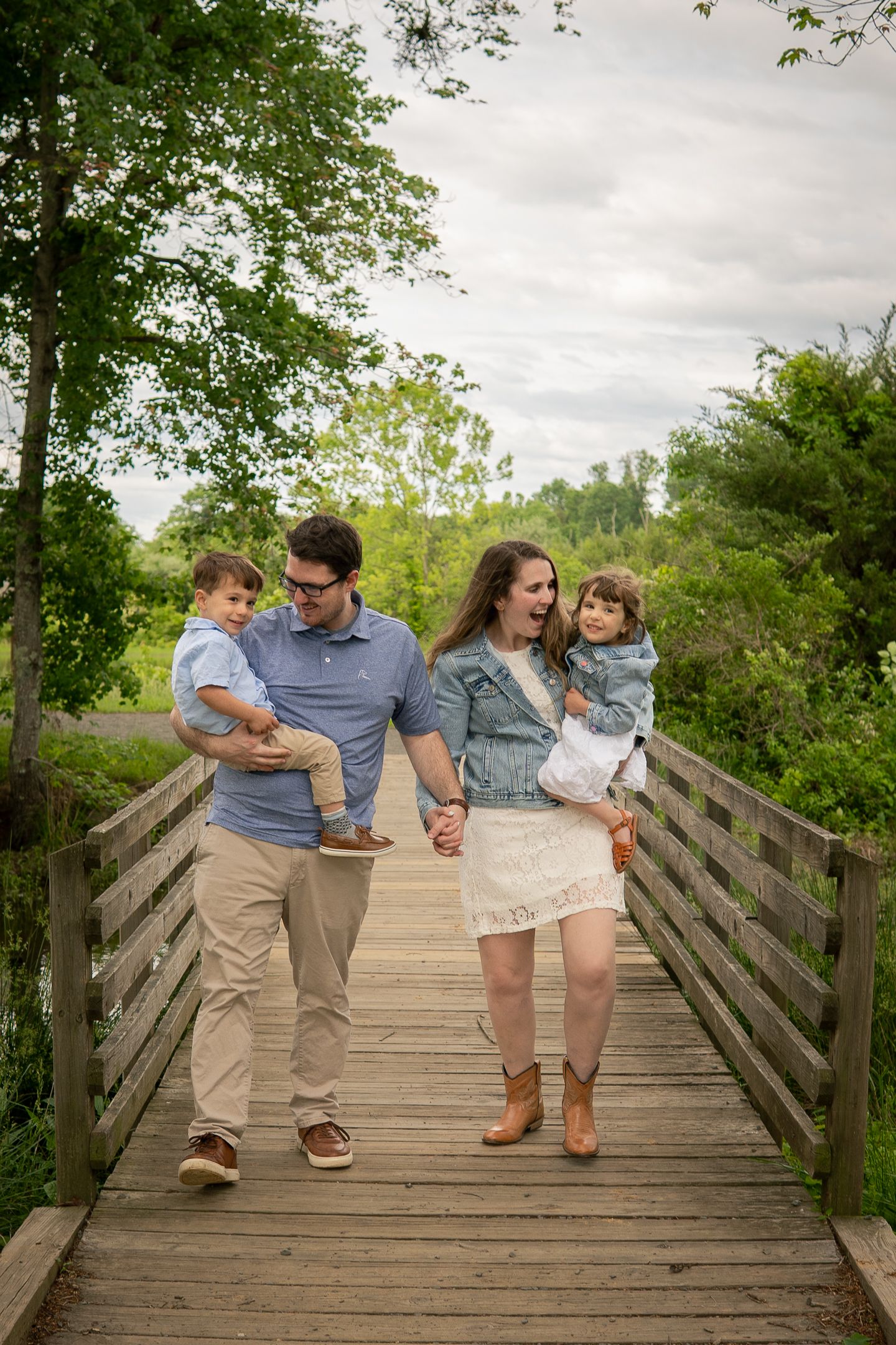 A couple walks with their children on a wooden bridge surrounded by lush green trees on a cloudy day.