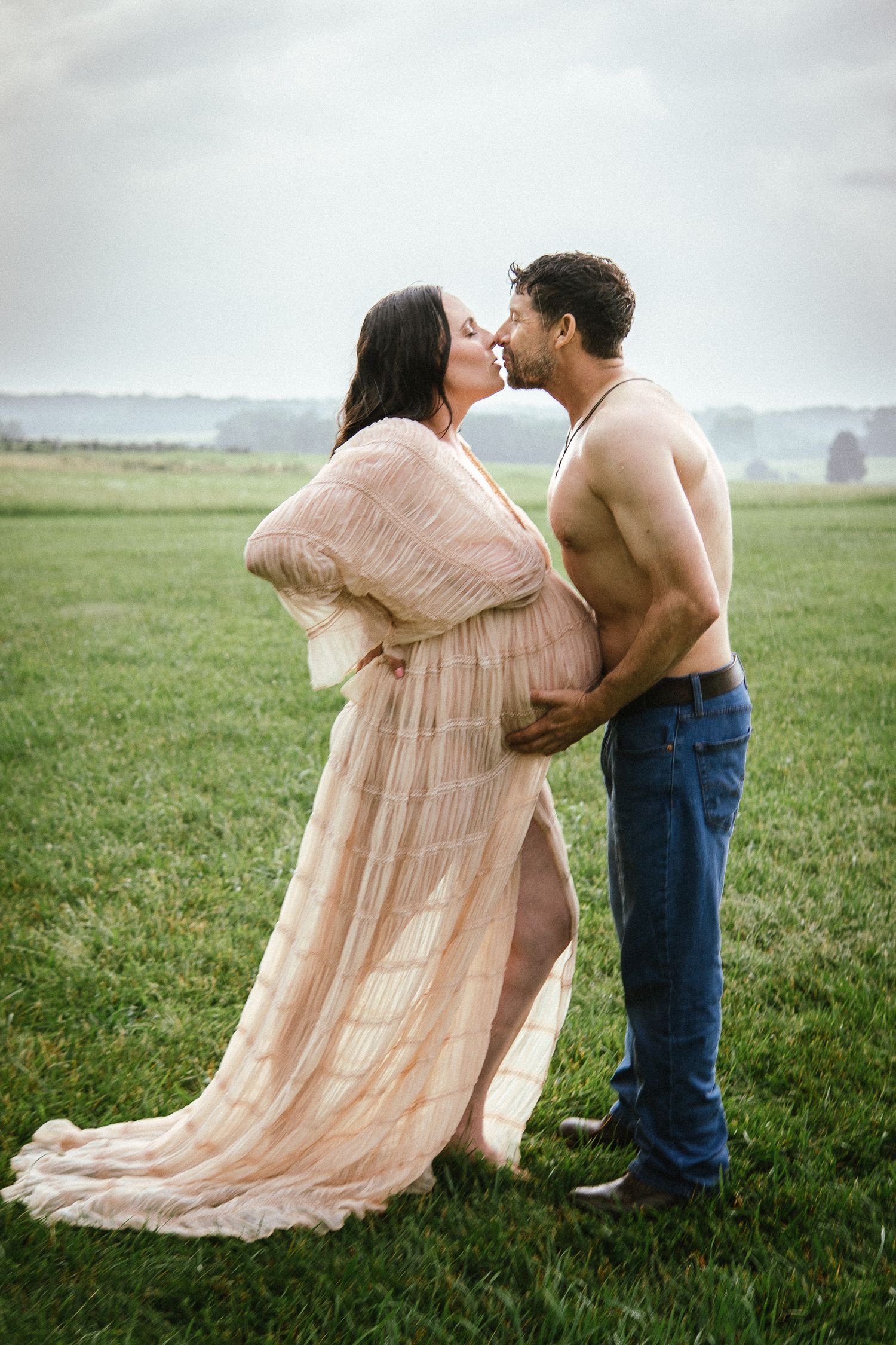 An intimate maternity portrait in a field features a couple embracing in soft peachy tones.