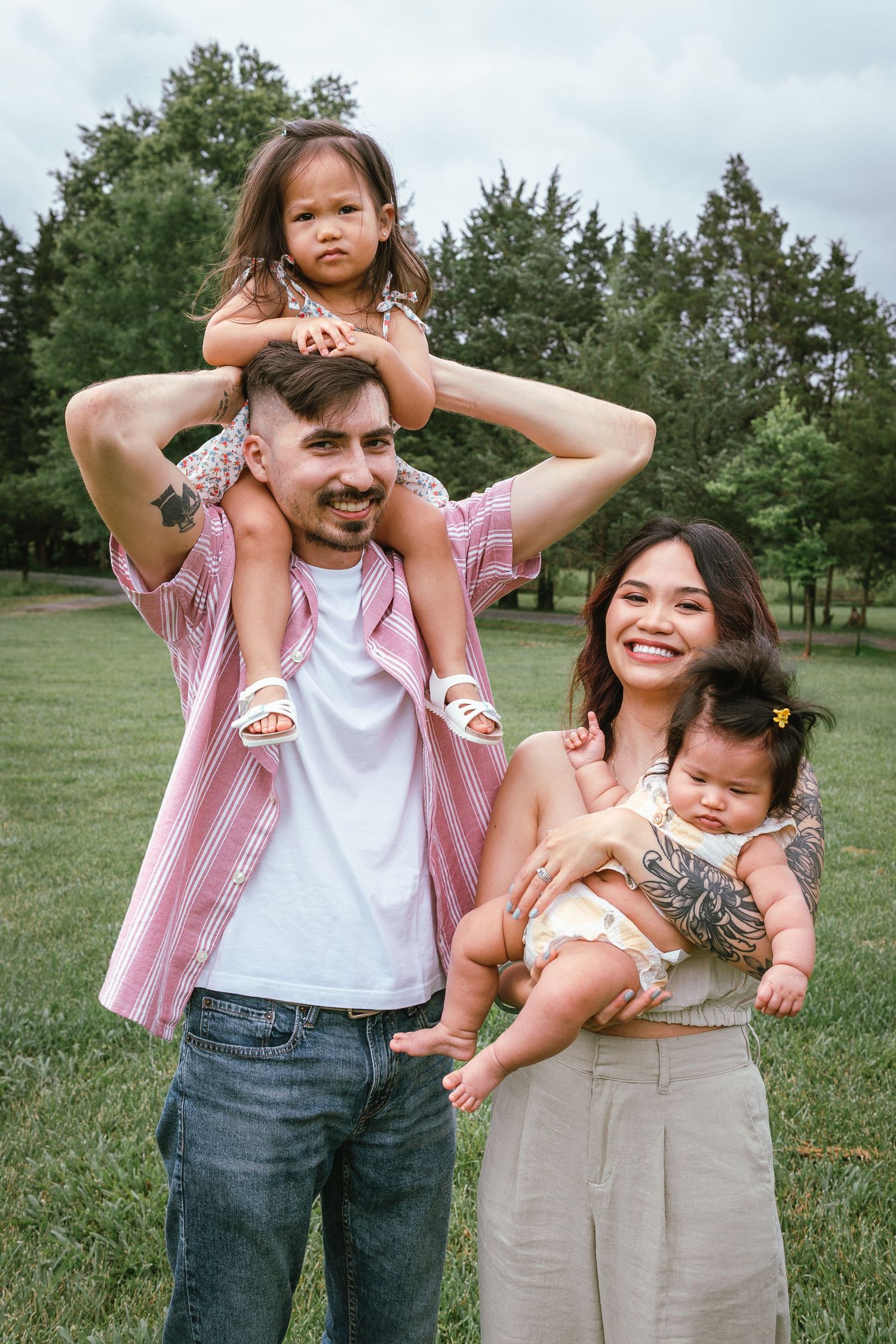 A family stands together on grass with green trees in the background, enjoying a casual outdoor moment.
