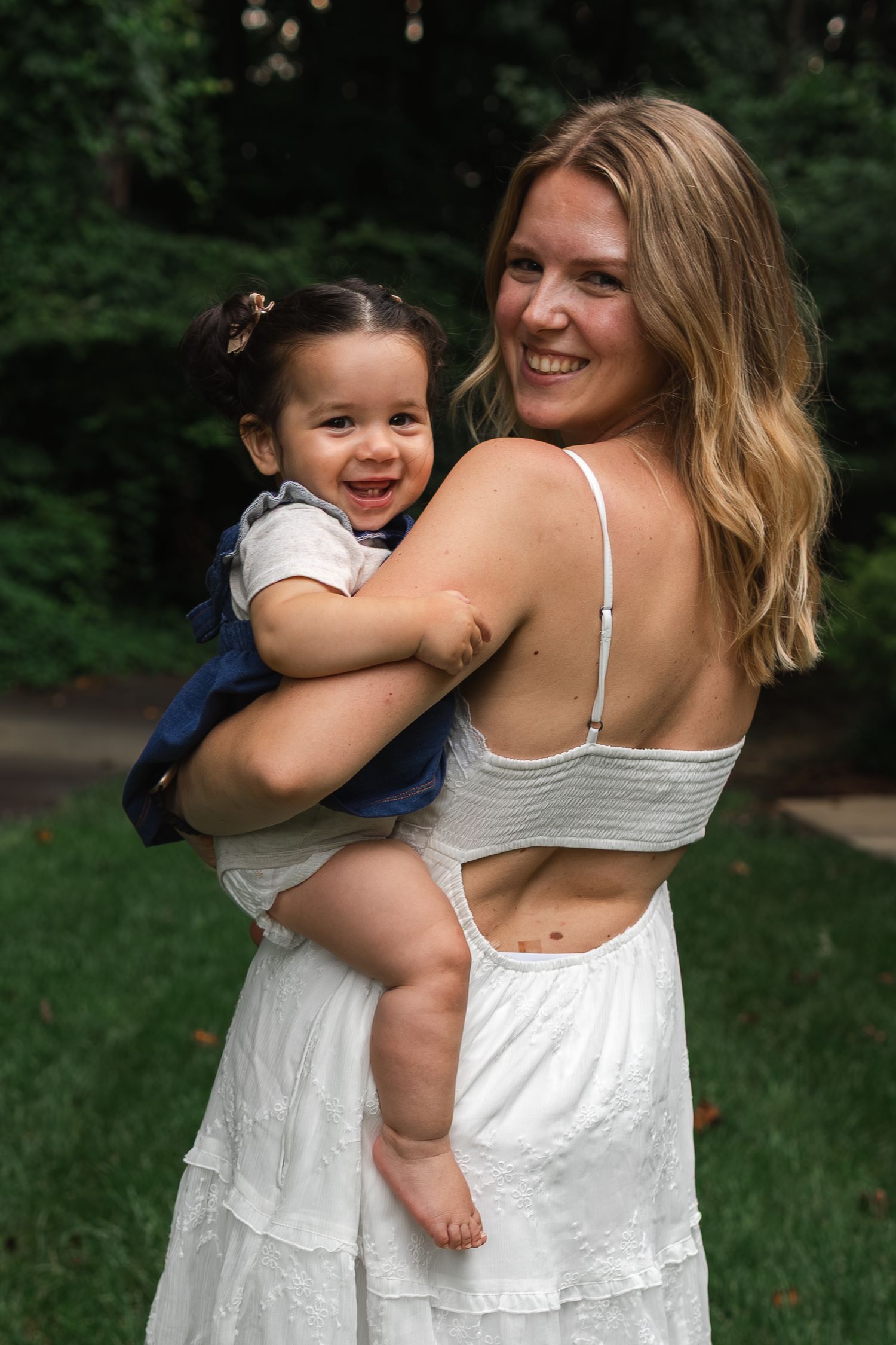 Woman in white summer dress holds smiling toddler outdoors in a garden setting.