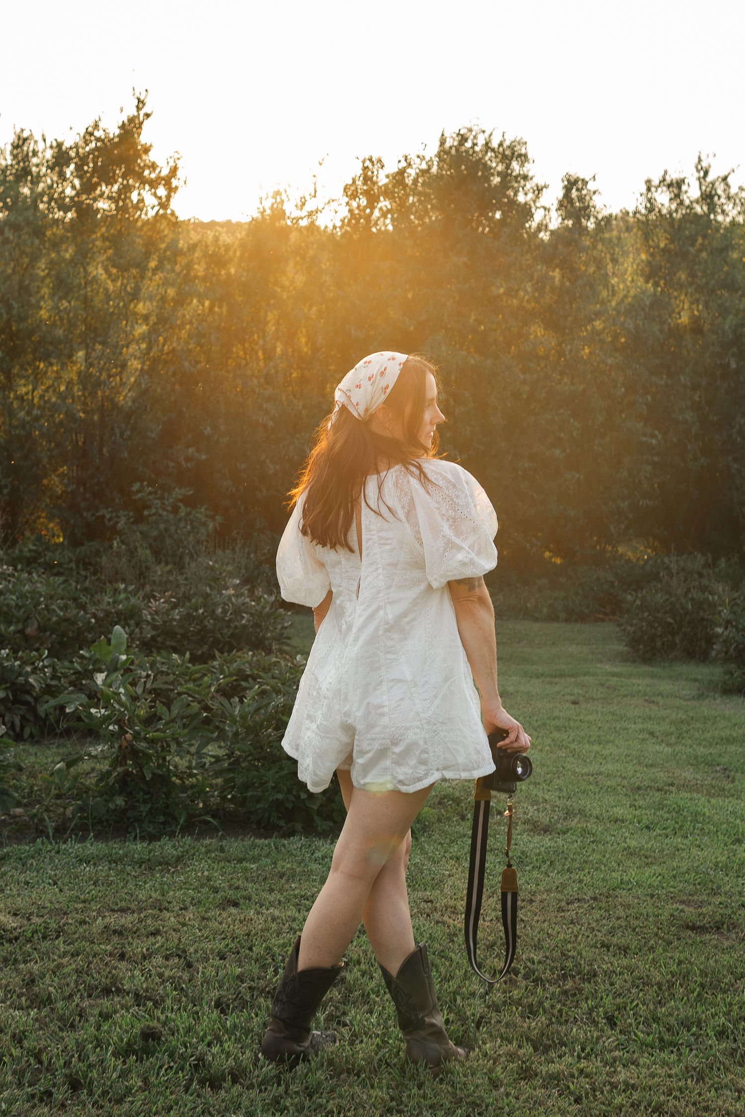 A person in a white flowy dress and boots stands in a meadow at sunset with golden light filtering through trees.