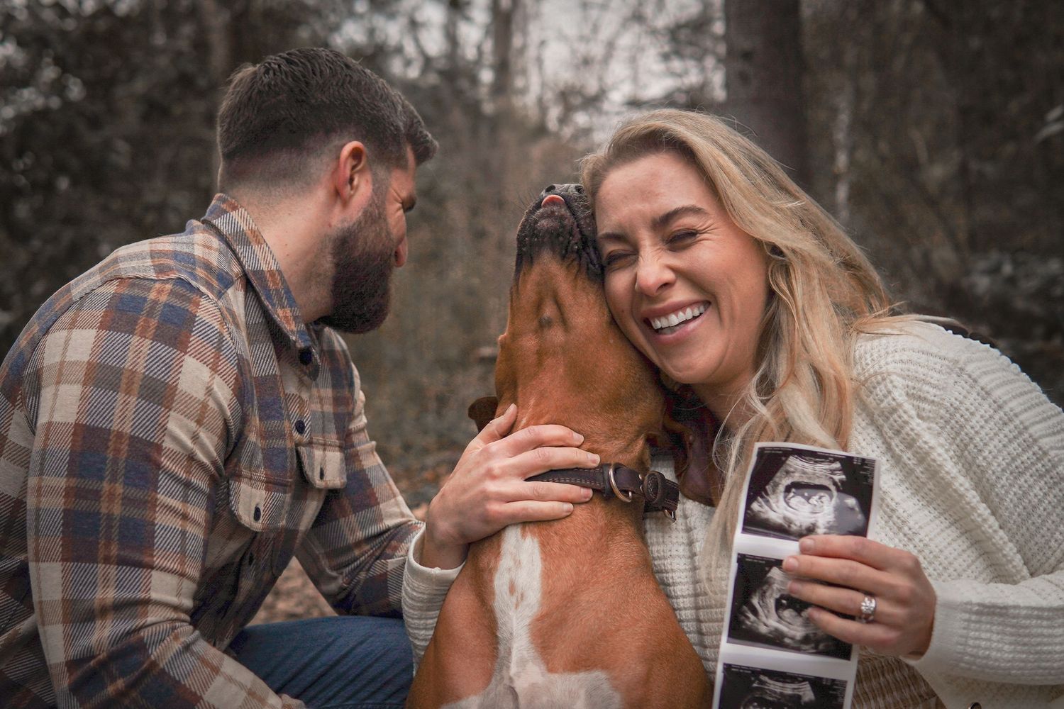 A joyful couple shares a laugh with their dog while displaying a sonogram photo in a wooded setting.