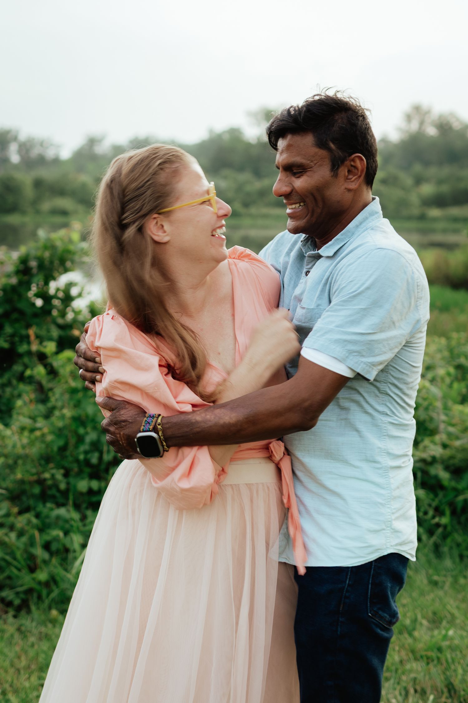 A couple shares a romantic embrace outdoors, with her wearing a flowing peach dress and him in a light blue shirt.