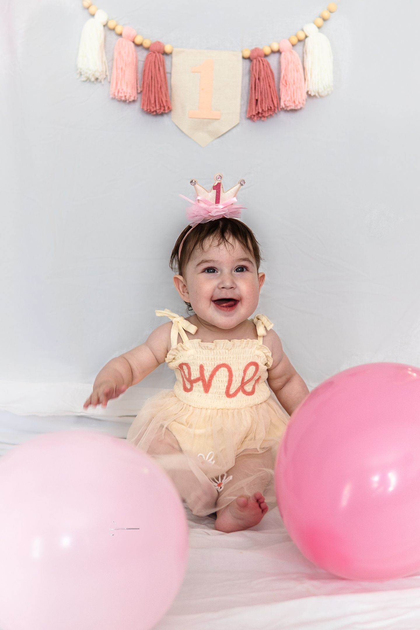 Baby in cream dress and pink crown smiles by pink balloons under tassel banner at first birthday photoshoot.