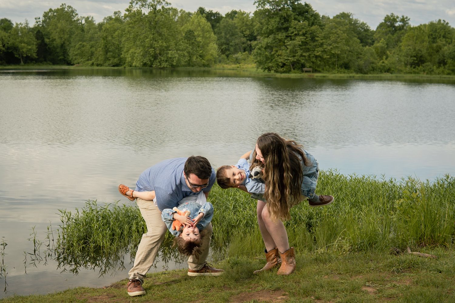 Two people kneel near the edge of a peaceful lake surrounded by green trees and grassy banks in late spring.