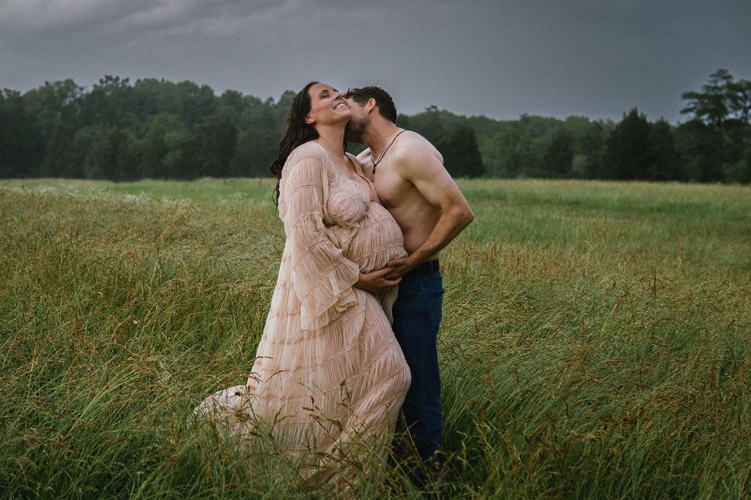 Two people in formal attire embrace in a scenic field at sunset creating a romantic outdoor portrait.