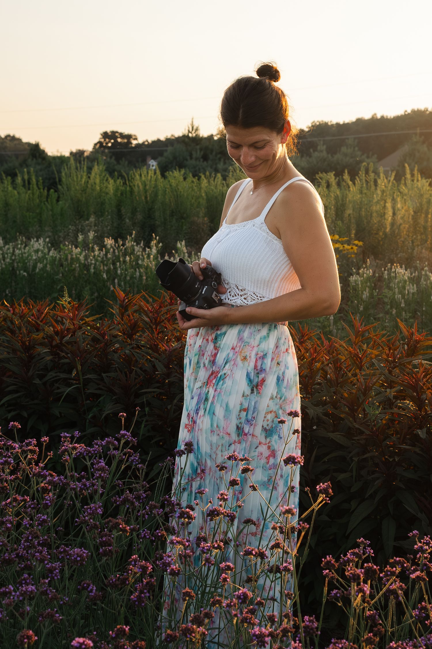 Photographer in floral dress looks down at camera while standing in beautiful flower garden at sunset.