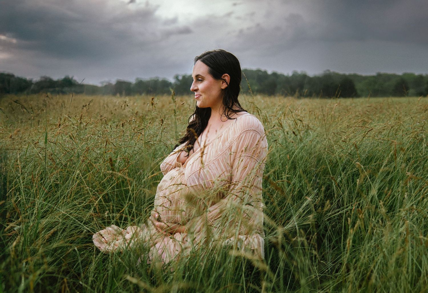 A woman in a light dress sits peacefully among tall grass with stormy skies overhead.