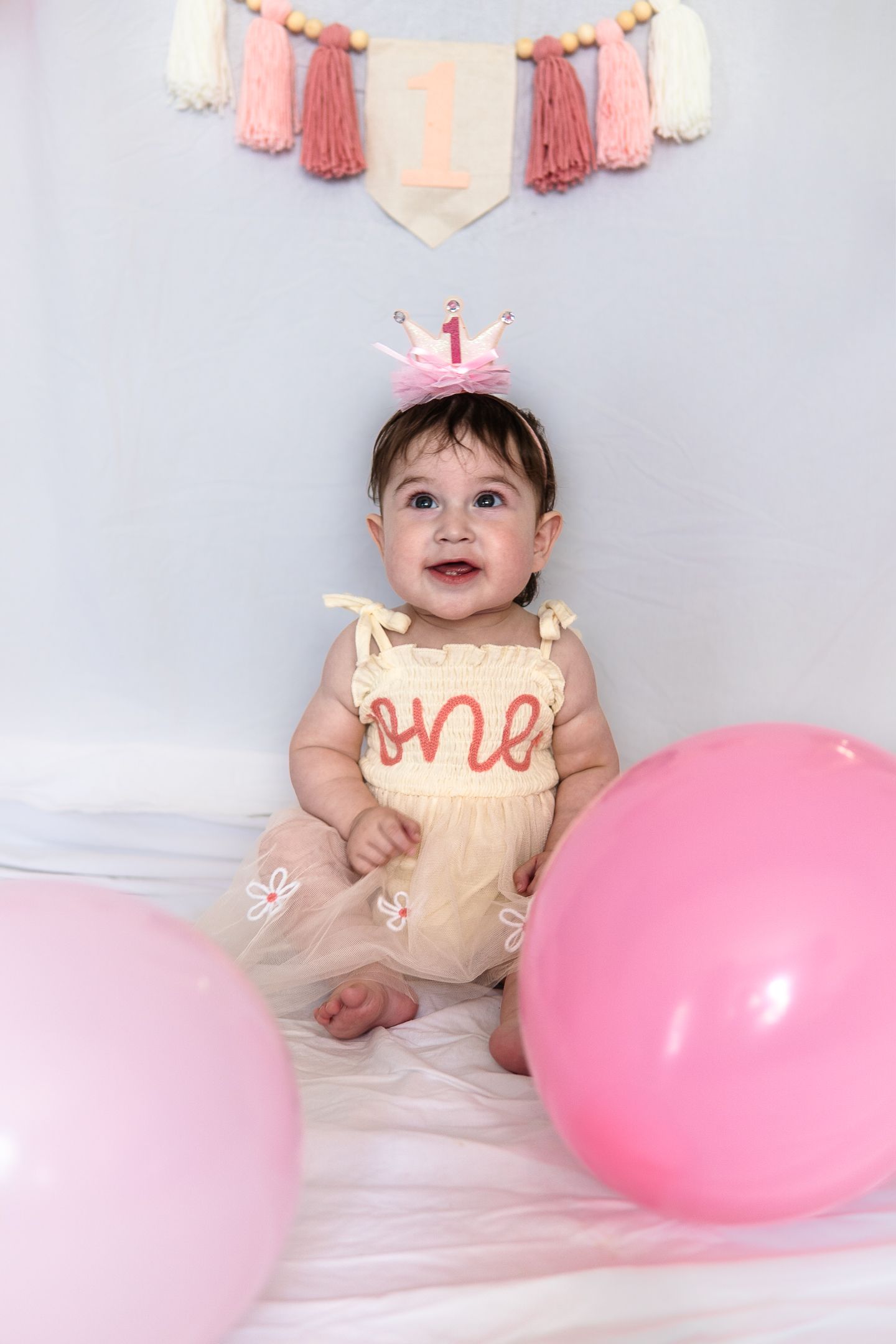 A baby wearing a peach tulle dress and crown sits on white fabric with pink balloons and tassel decor in background.