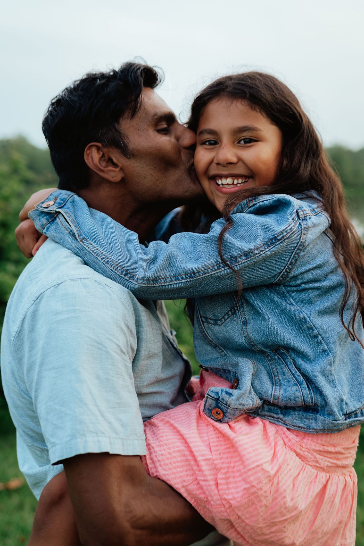 A playful couple embraces and laughs together outdoors, with one wearing a denim jacket and coral dress.