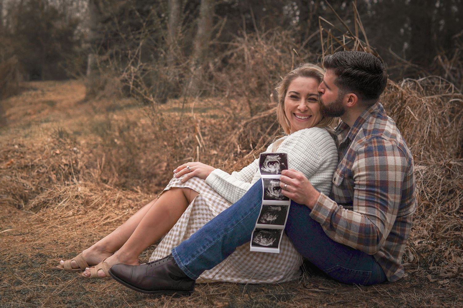 Couple sitting on ground outdoors holding ultrasound images during pregnancy announcement photoshoot in autumn setting.