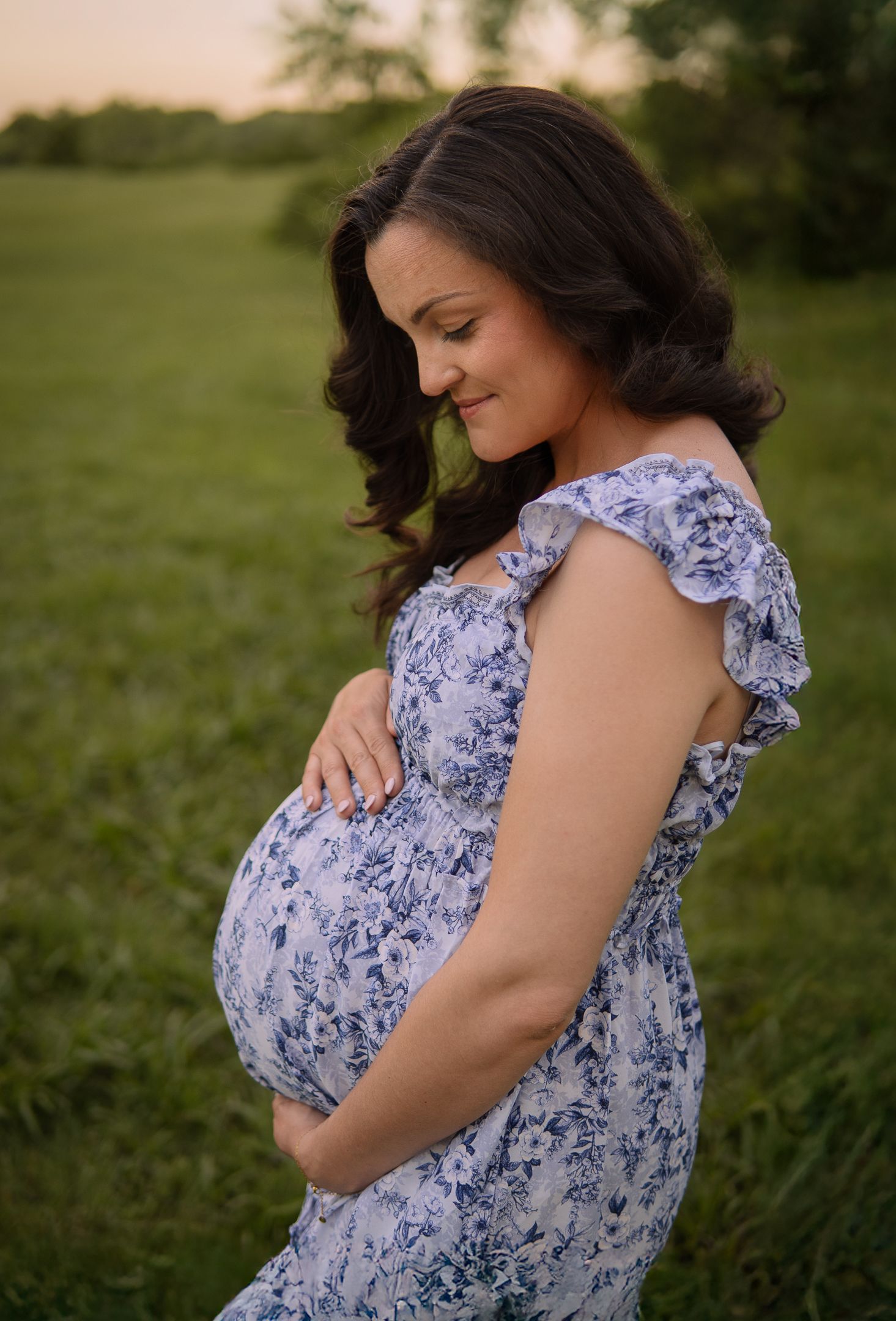 A pregnant woman in a blue and white floral dress stands in a grassy field at sunset, gently touching her baby bump.