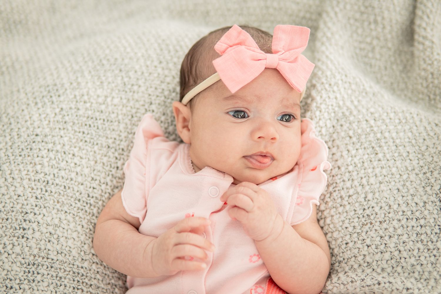 A baby wearing a pink outfit and matching bow headband lies on a soft grey blanket.