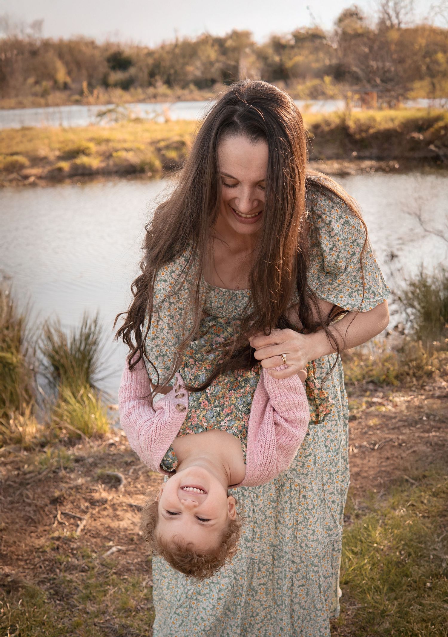 Woman in floral dress holds and playfully tips back small child by riverbank during golden hour at sunset.