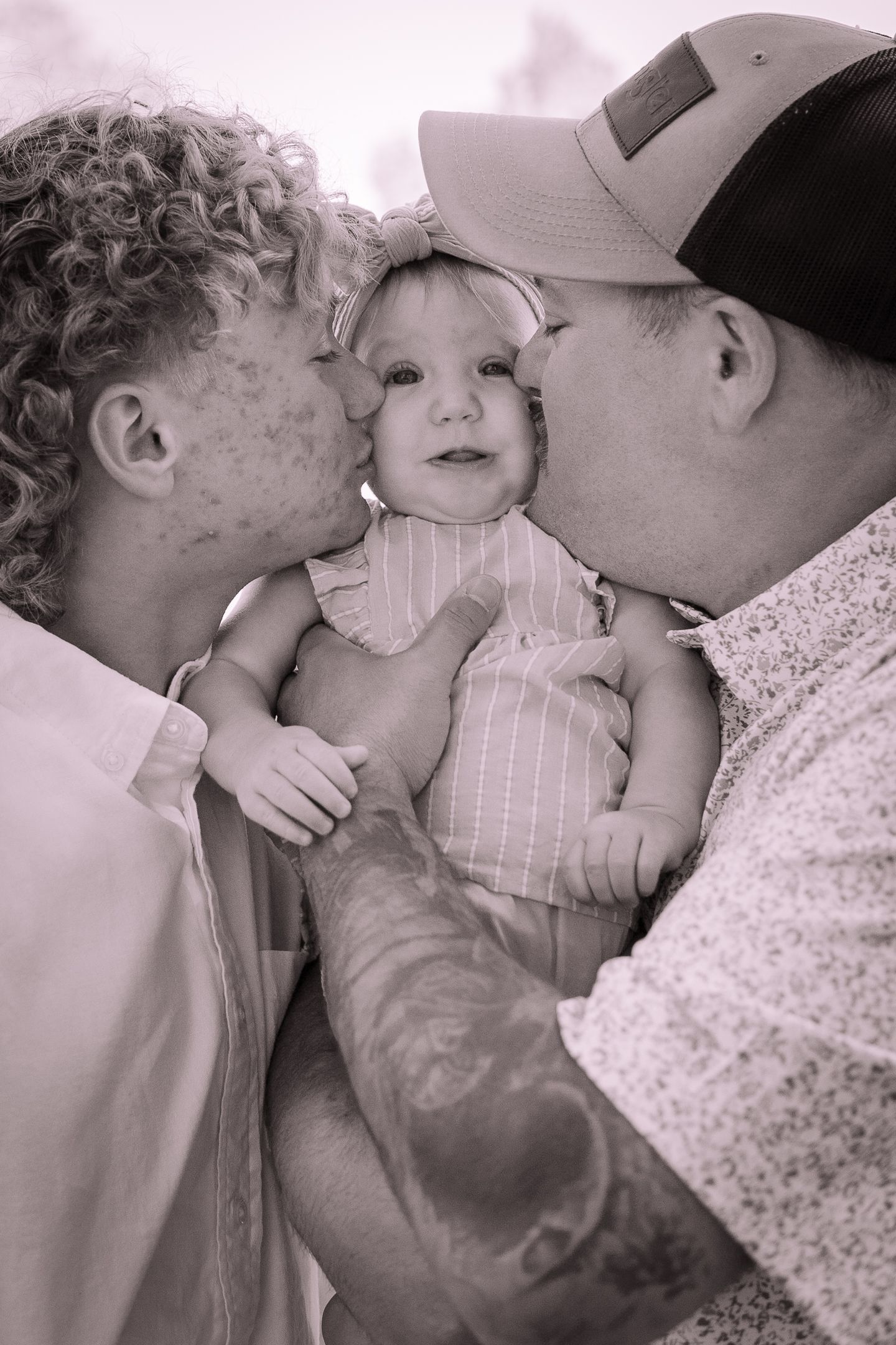 A heartwarming black and white photo of two people kissing a baby in a pinstriped outfit.