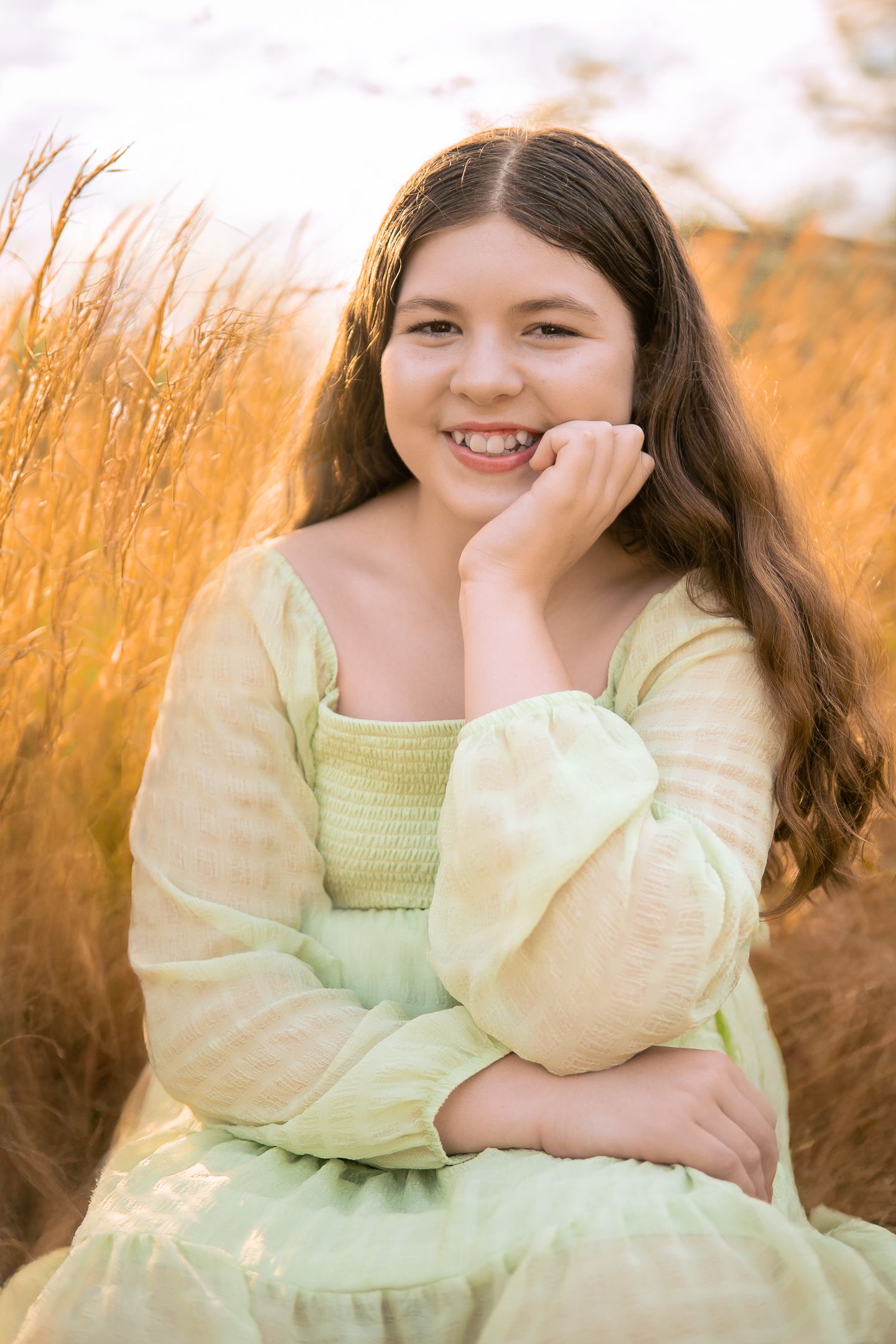 A joyful portrait in a yellow spring dress sitting among golden wheat at sunset with warm glowing light.