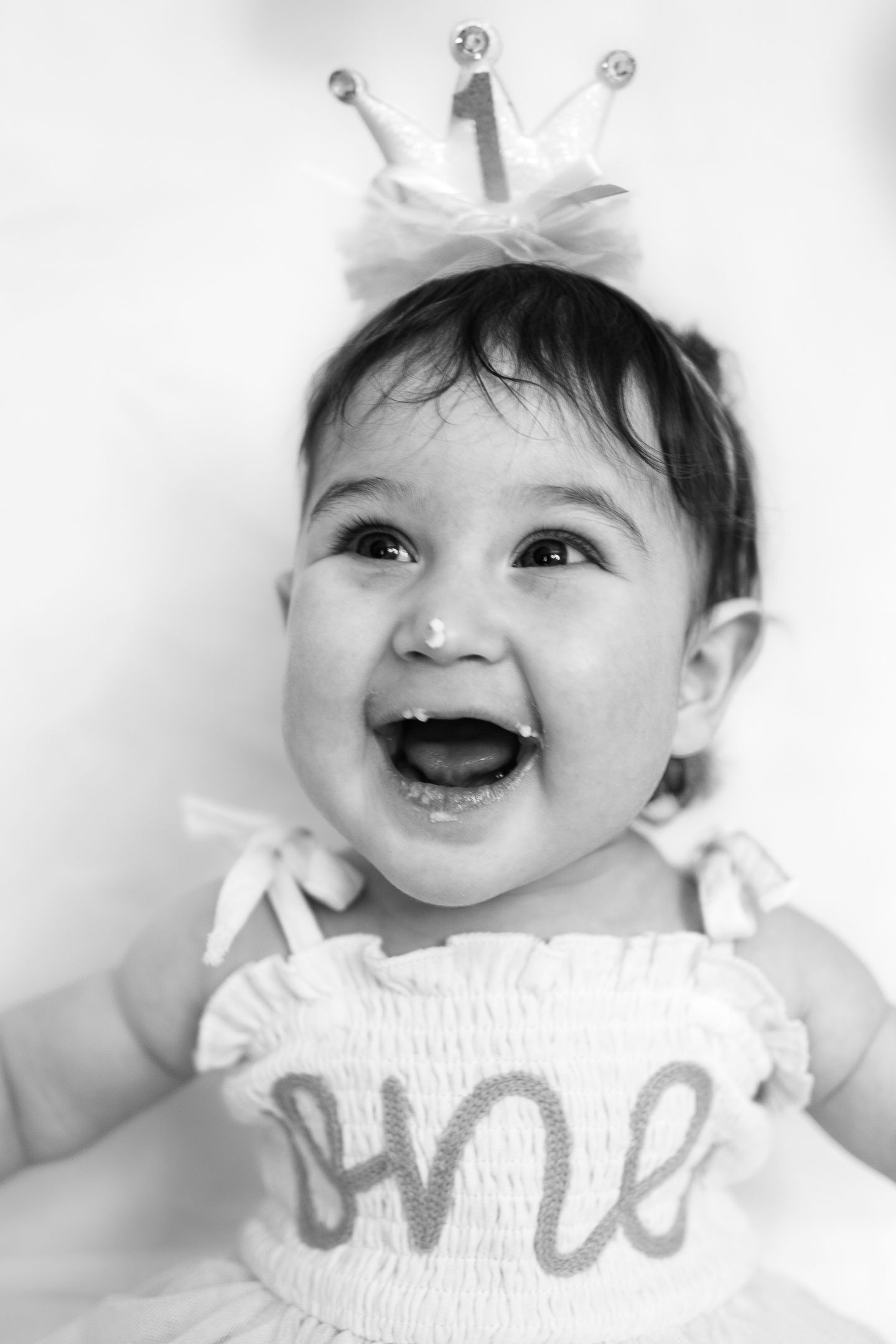 Black and white portrait of a baby wearing a white dress with 'one' text and a small crown, laughing joyfully.