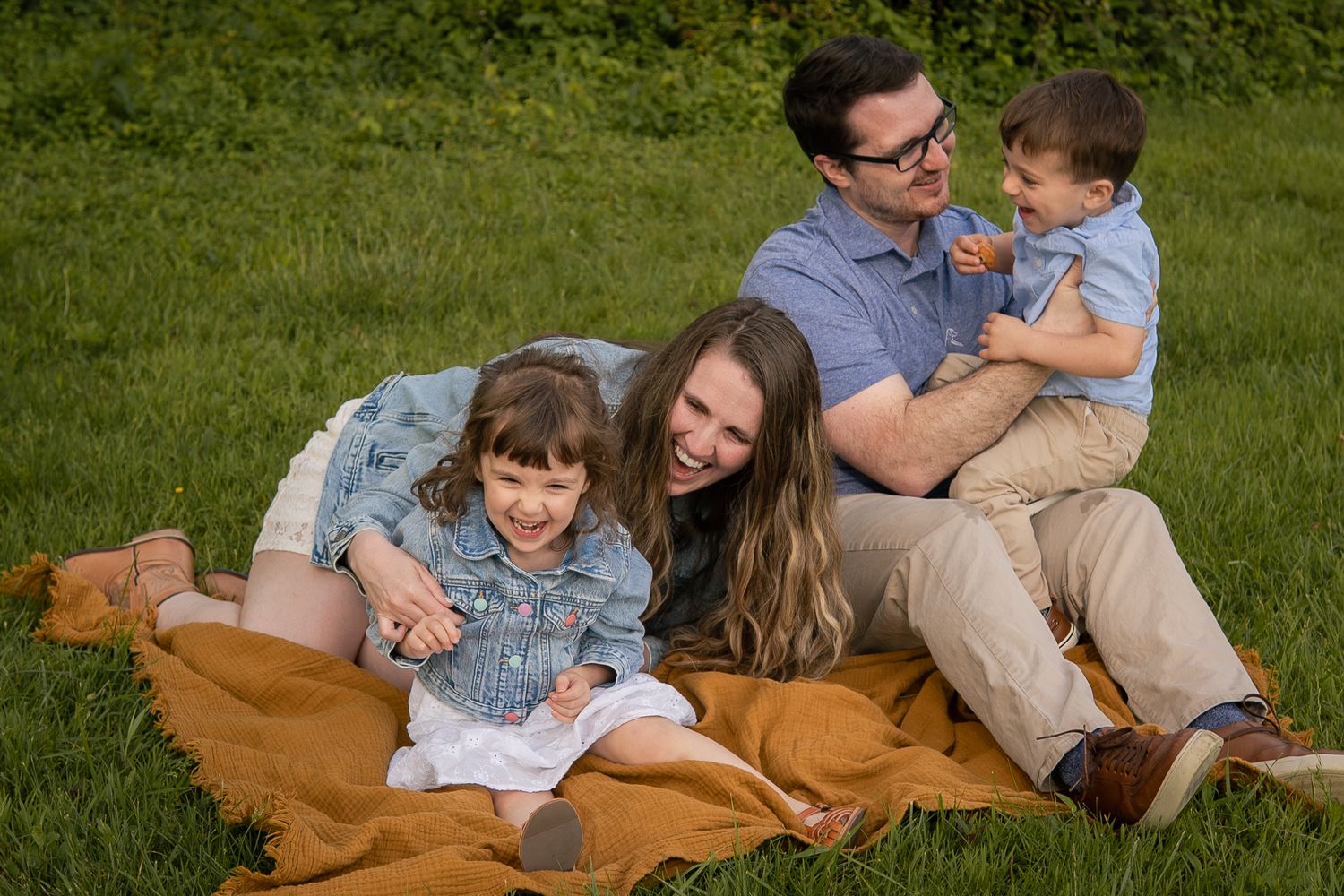 A cheerful family sits together on a brown blanket in a sunny green lawn during an outdoor photo session.