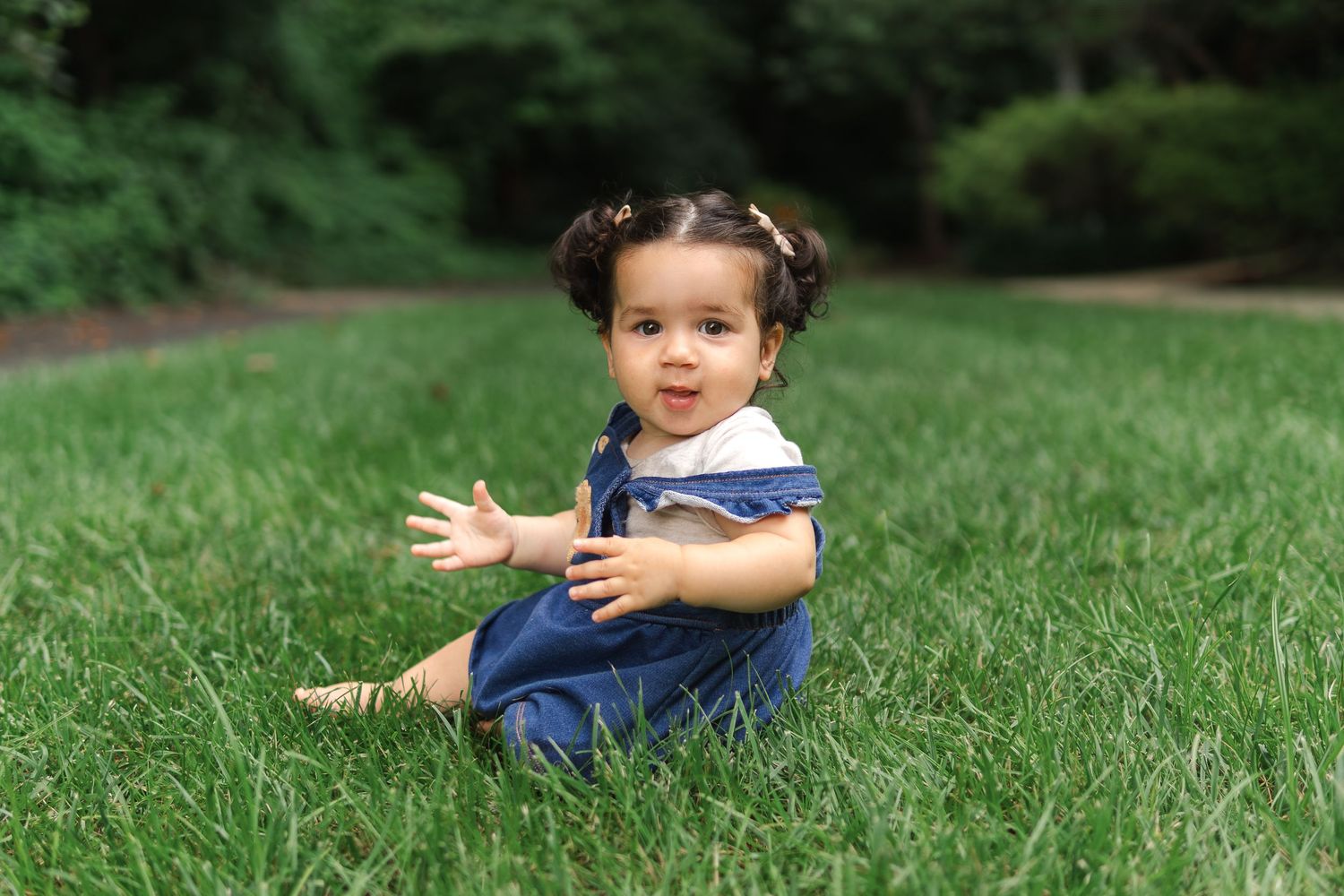 A small child in denim overalls sits on lush green grass with trees in the background.