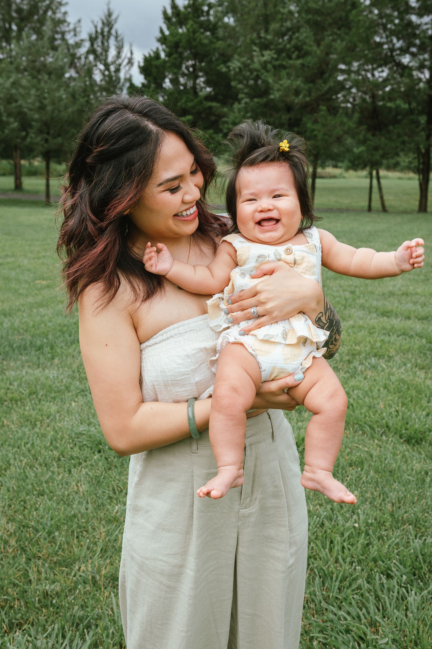 A joyful outdoor photoshoot in a grassy field shows playful moments between a parent and baby in matching beige outfits.