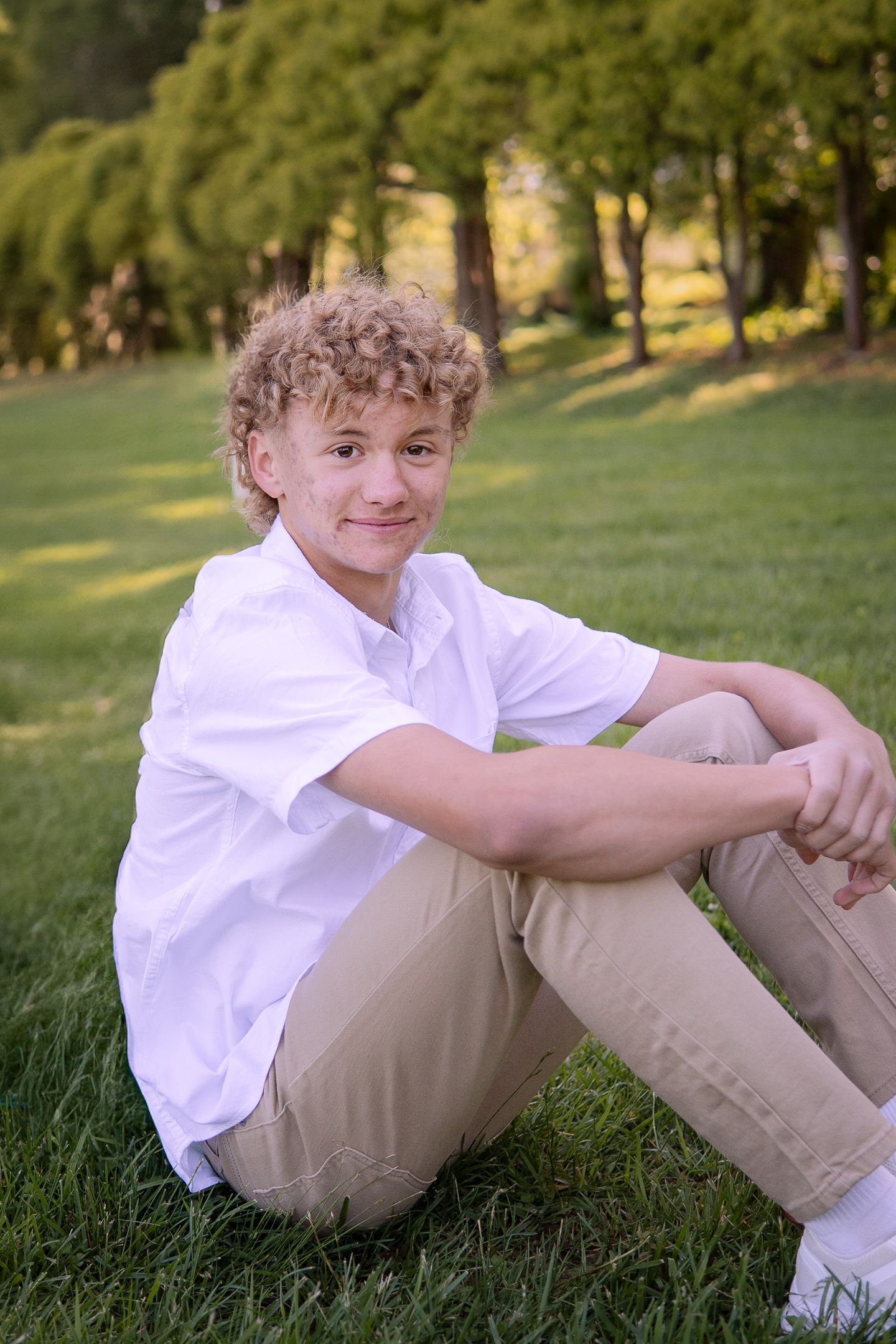 A casual outdoor portrait of someone sitting on grass wearing white shirt and khaki pants.