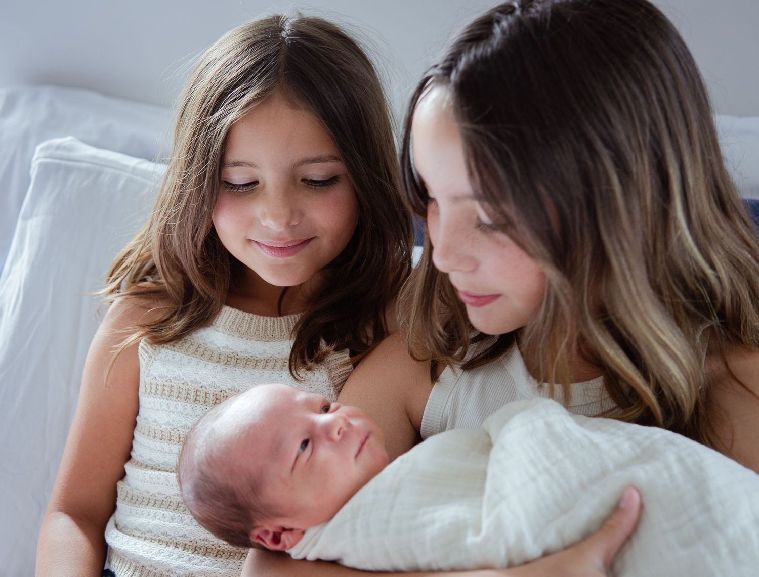 Family moment with siblings gazing lovingly at newborn baby in white wrap.